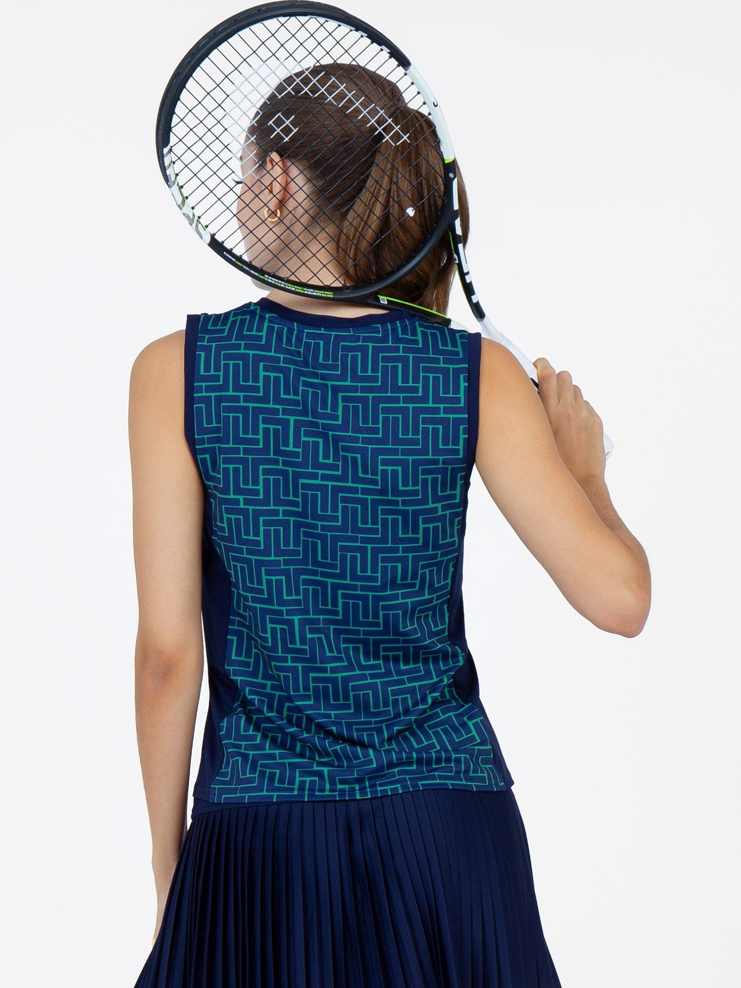 A woman faces away, holding a tennis racket over her right shoulder. She wears the inPhorm NYC Harper Performance Tank in Ink Tessel/Ink and a navy pleated skirt, offering moisture-wicking comfort against a plain white background.