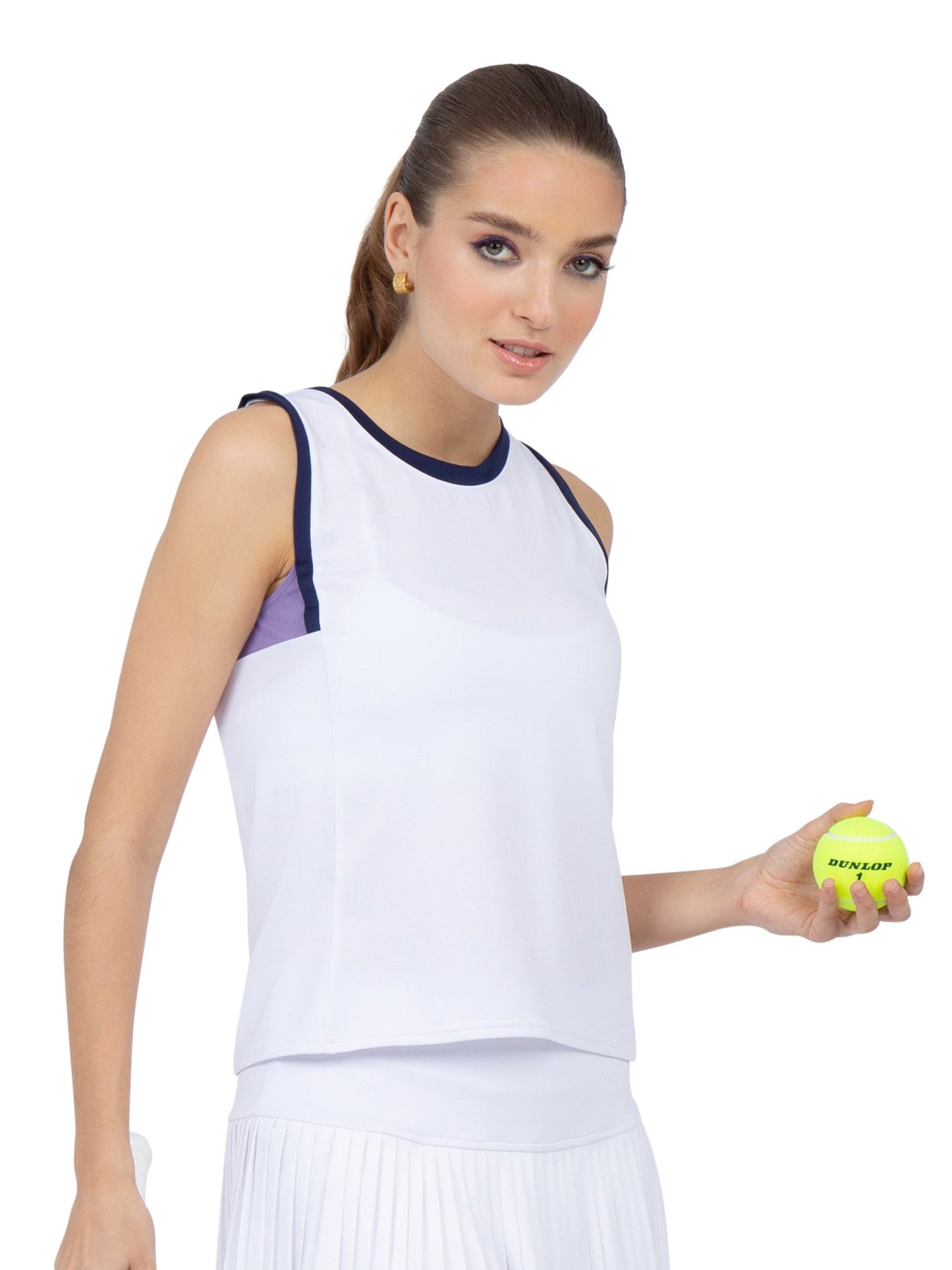 A woman with long brown hair tied back wears the inPhorm NYC Harper Performance Tank in White/Lavender Combo and a pleated skirt. Holding a yellow tennis ball, she looks to the side, ready for action in stylish UPF sun protection.