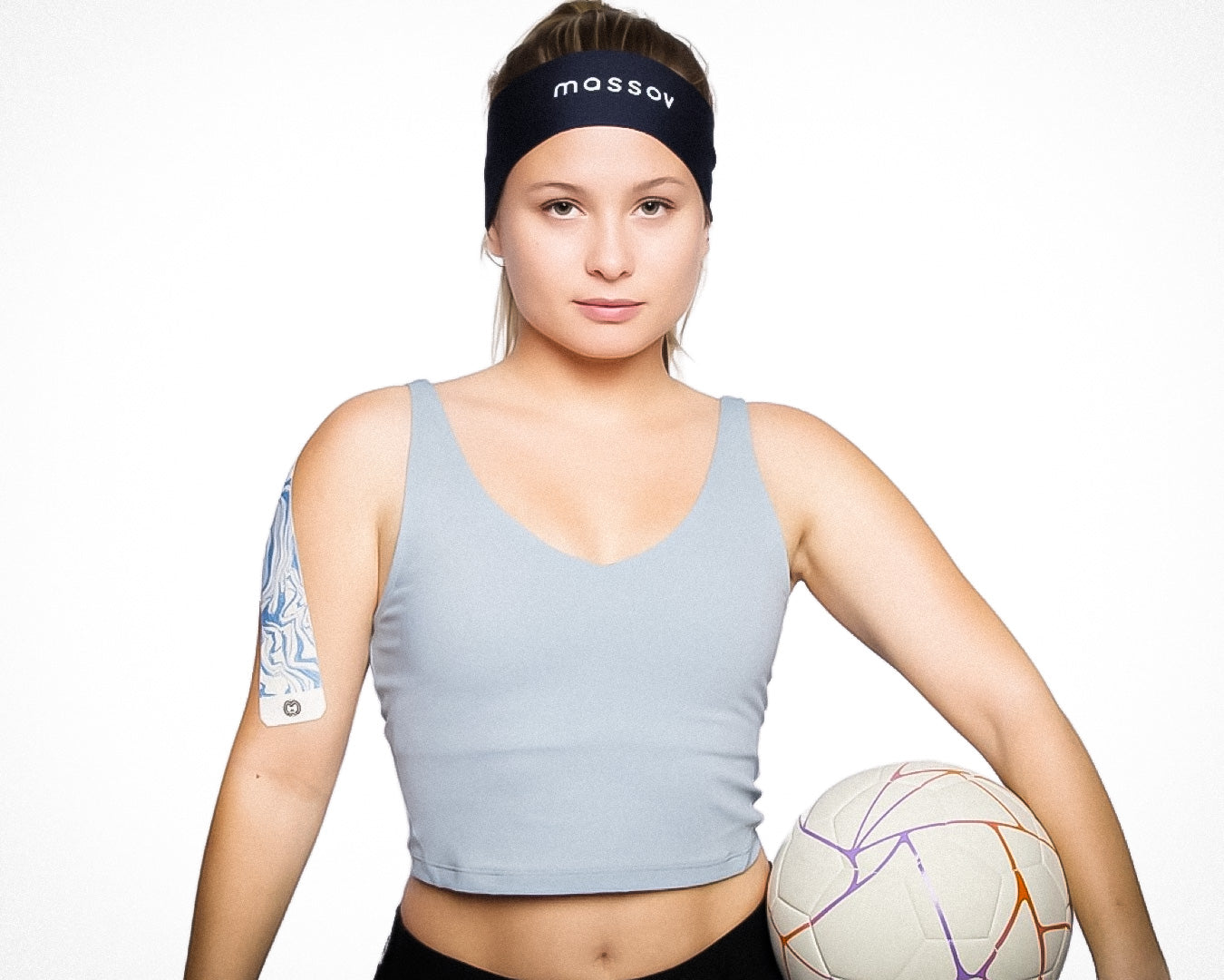 A woman wearing a light blue tank top and the Massov Headband Tie stands confidently holding a soccer ball. She has a patterned adhesive patch on her upper arm, with a neutral expression against a plain white background.