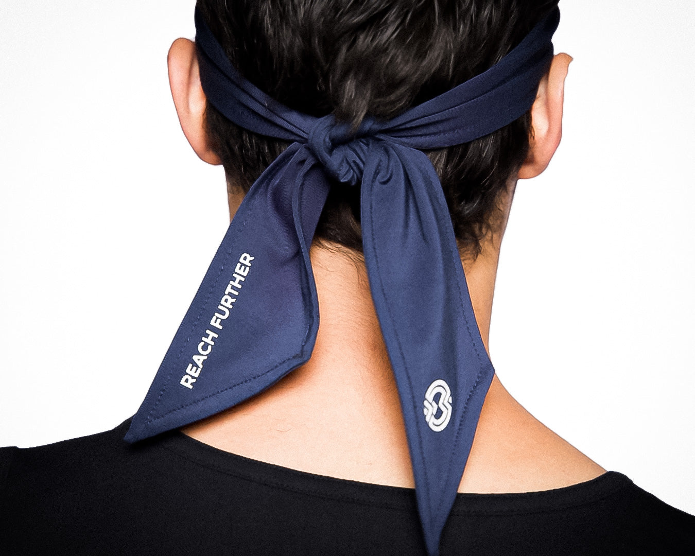 A person seen from behind wears a navy blue Massov Headband Tie with “REACH FURTHER” in white on one tail and a small circular logo on the other. The sweat-wicking bandana is paired with a black shirt.