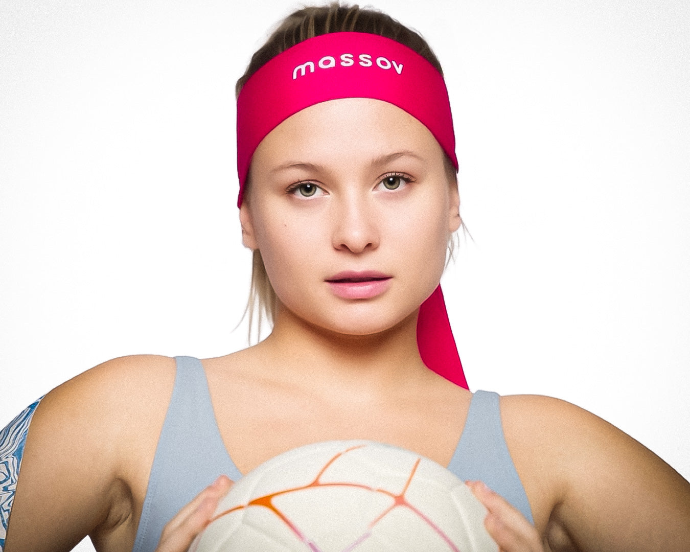 A young woman with light skin and straight blonde hair wears a bright pink Massov Headband Tie, looks forward with a neutral expression, holds a white soccer ball with orange lines, and wears a pale blue athletic top.