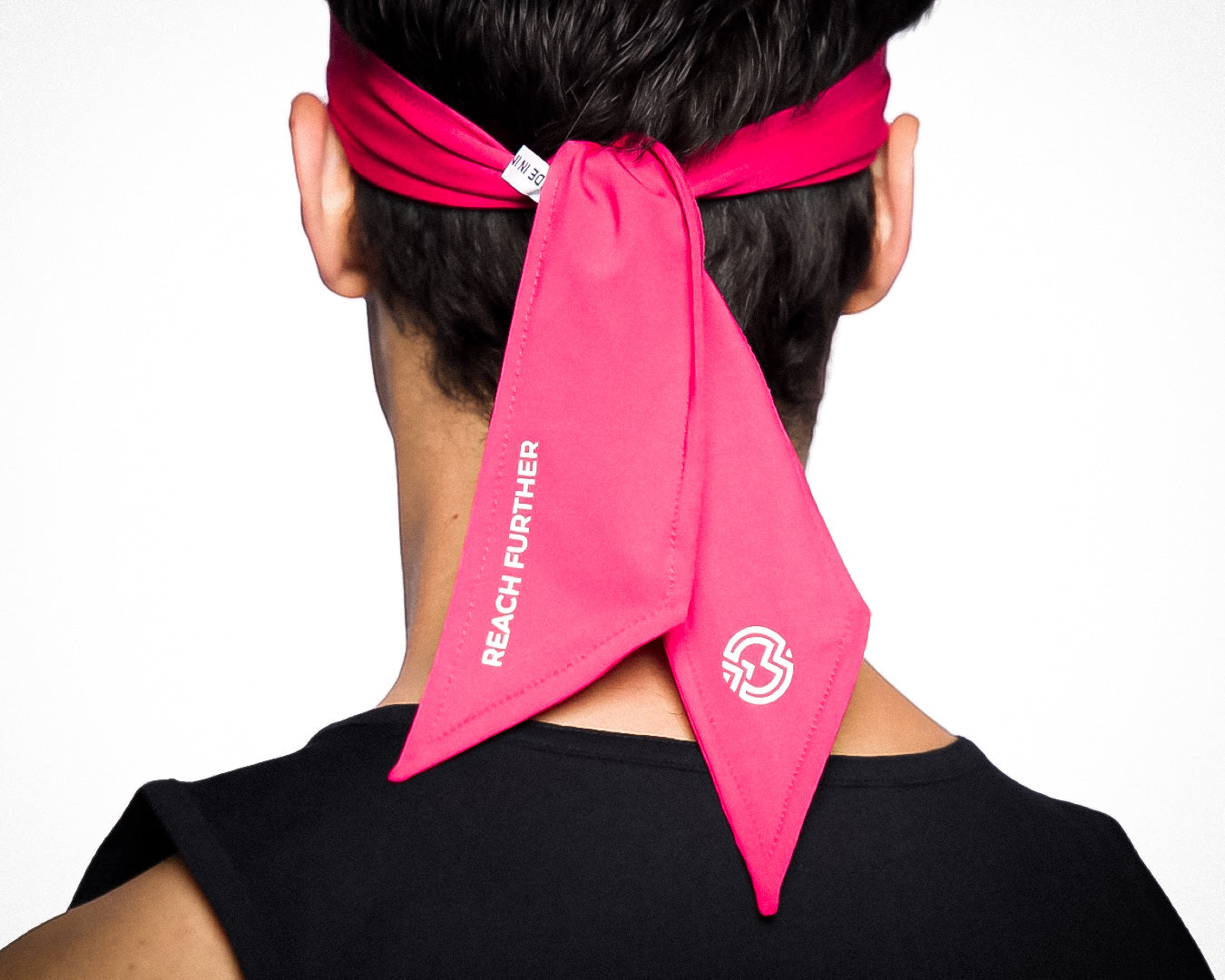 A person seen from behind wears a bright pink Massov Headband Tie, featuring white “REACH FURTHER” text and logo. The headband is sweat-wicking and tied at the back. The person has on a sleeveless black top against a white background.