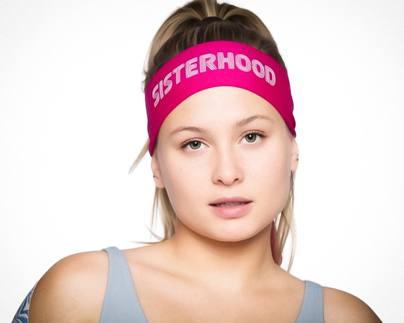 A young woman with light skin and long blonde hair tied back wears a pink Massov Headband Tie reading SISTERHOOD in bold white letters. She smiles slightly at the camera, wearing a light blue tank top against a plain white background.