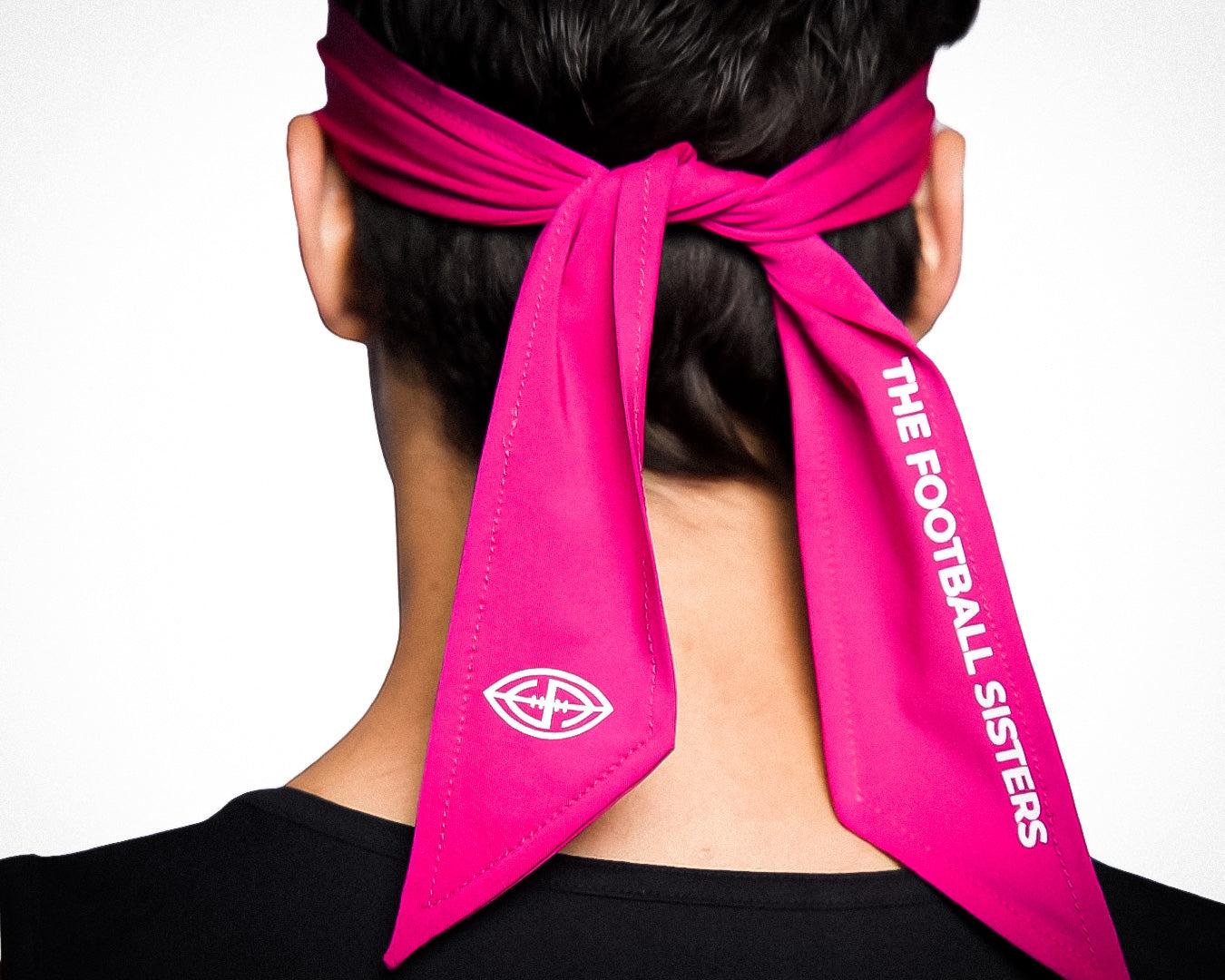 Seen from behind, a person wears the Massov Headband Tie in bright pink, featuring "THE FOOTBALL SISTERS" in white on one tail and a small white geometric logo on the other, set against a plain white background.