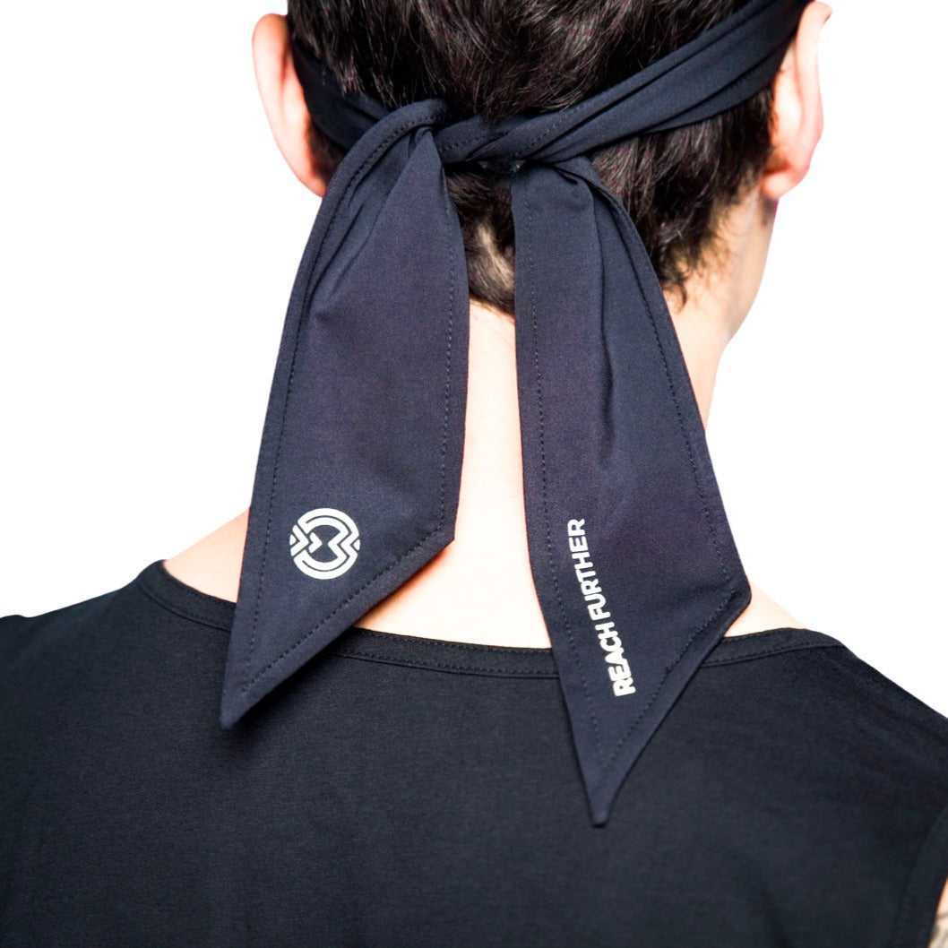A person in a black top is seen from behind wearing a Massov Headband Tie in black, tied at the back with two long ends—one displaying a geometric logo and the other reading “REACH FURTHER” in white.