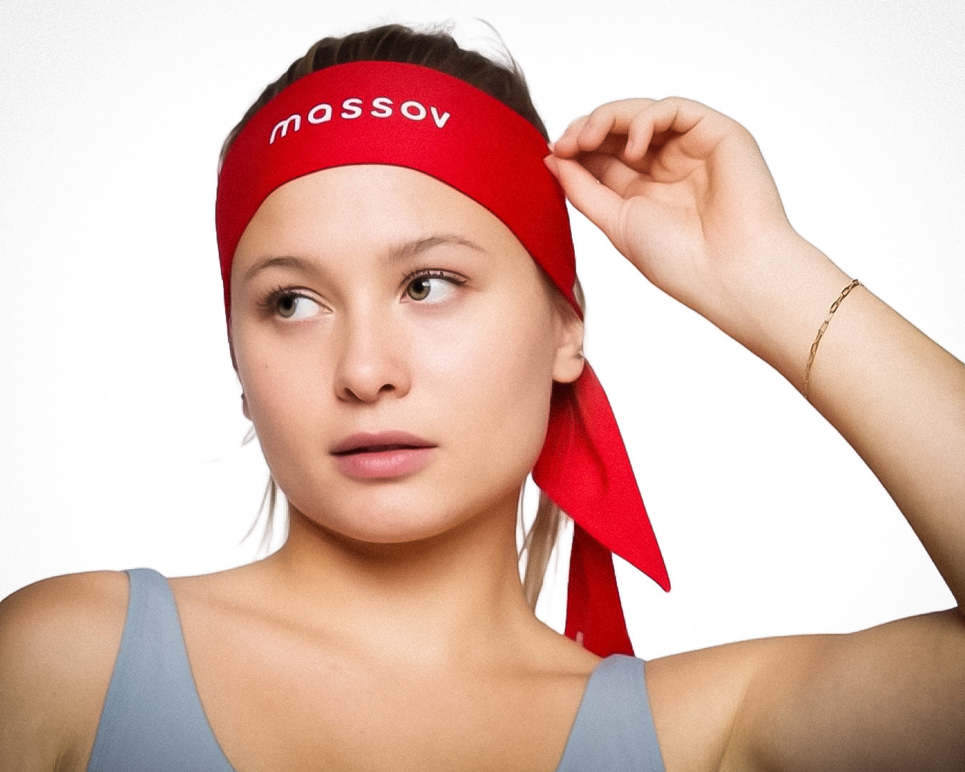 A young woman with light skin and straight hair pulled back wears the Massov Headband Tie in red, featuring the brand's name. She pairs it with a pale blue tank top, a delicate gold bracelet, and stands against a plain white background.