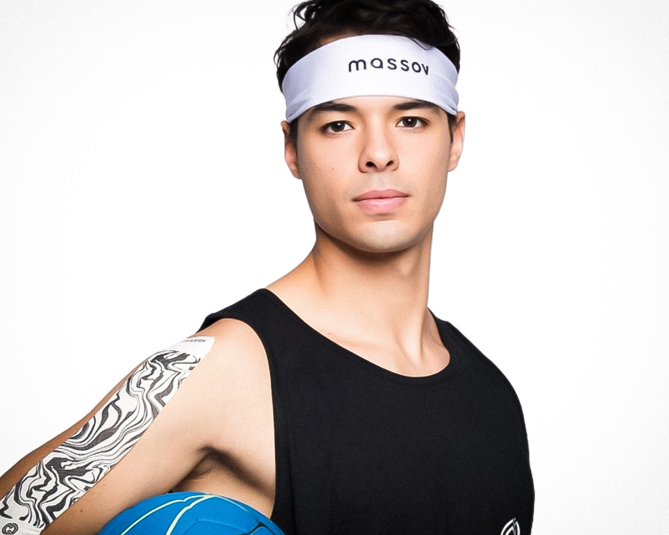 A young man wears a white Massov Headband Tie and black tank top, holding a blue volleyball. Black-and-white patterned tape is on his right shoulder as he looks confidently at the camera against a plain white background.