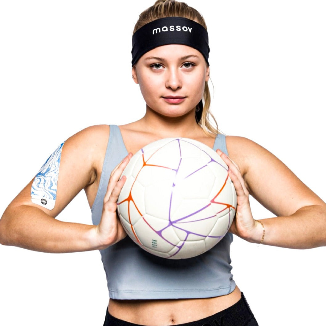 A confident young woman wears a Massov Headband Tie in black and a blue tank top, holding a white soccer ball with orange and purple lines. Patterned athletic tape is on her right upper arm, standing ready against a white background.