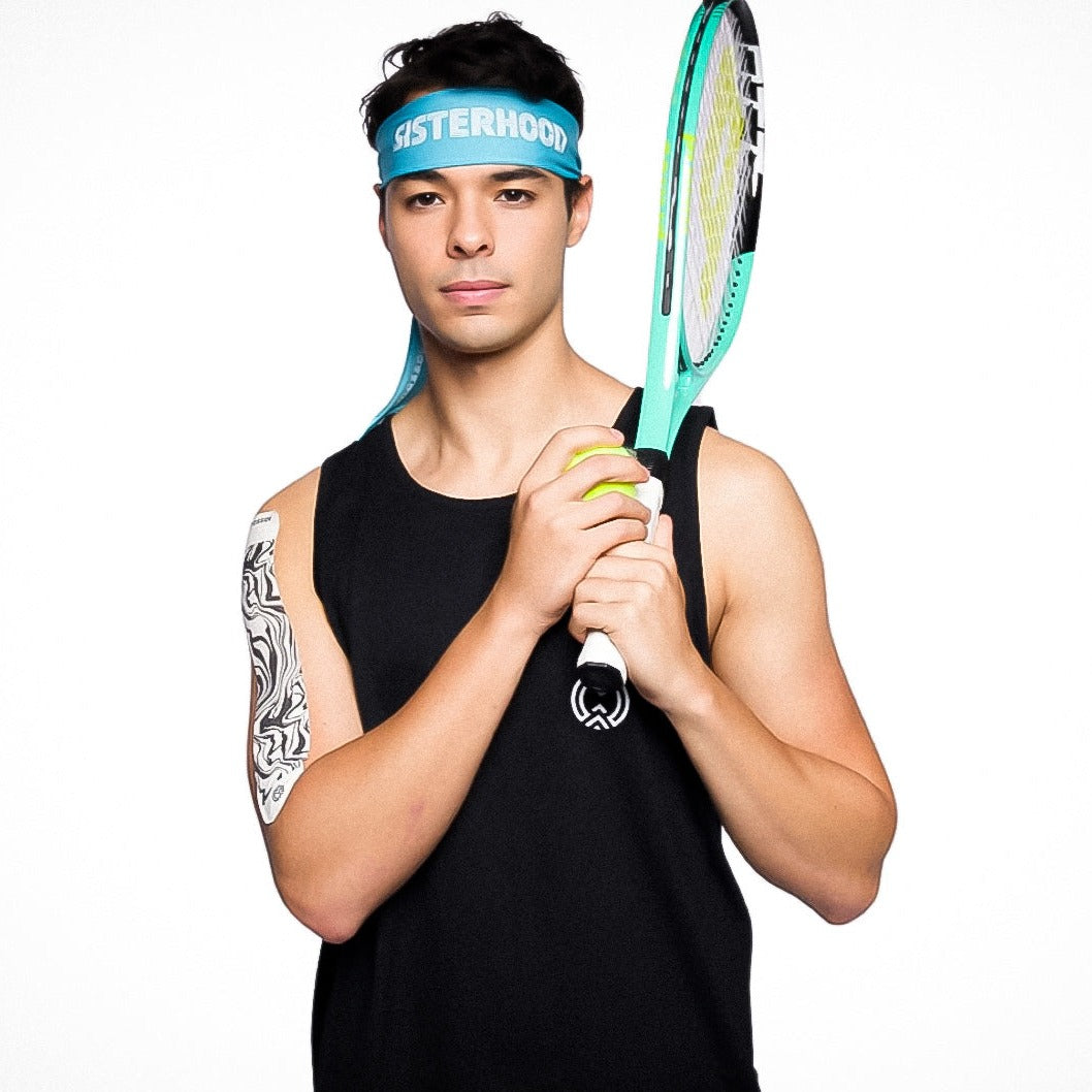 A young man in a black tank top holds a tennis racquet and ball. He sports the Massov Headband Tie, a blue sweat-wicking accessory labeled “SISTERHOOD,” and stands confidently against a plain white background, showing an abstract tattoo.