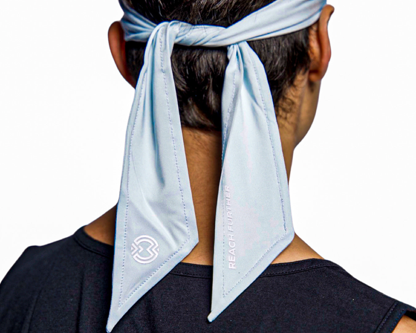 A person wearing a black shirt is seen from behind with a Massov Headband Tie in light blue, featuring two long pointed ends. One end displays "REACH FURTHER" and an abstract white logo.