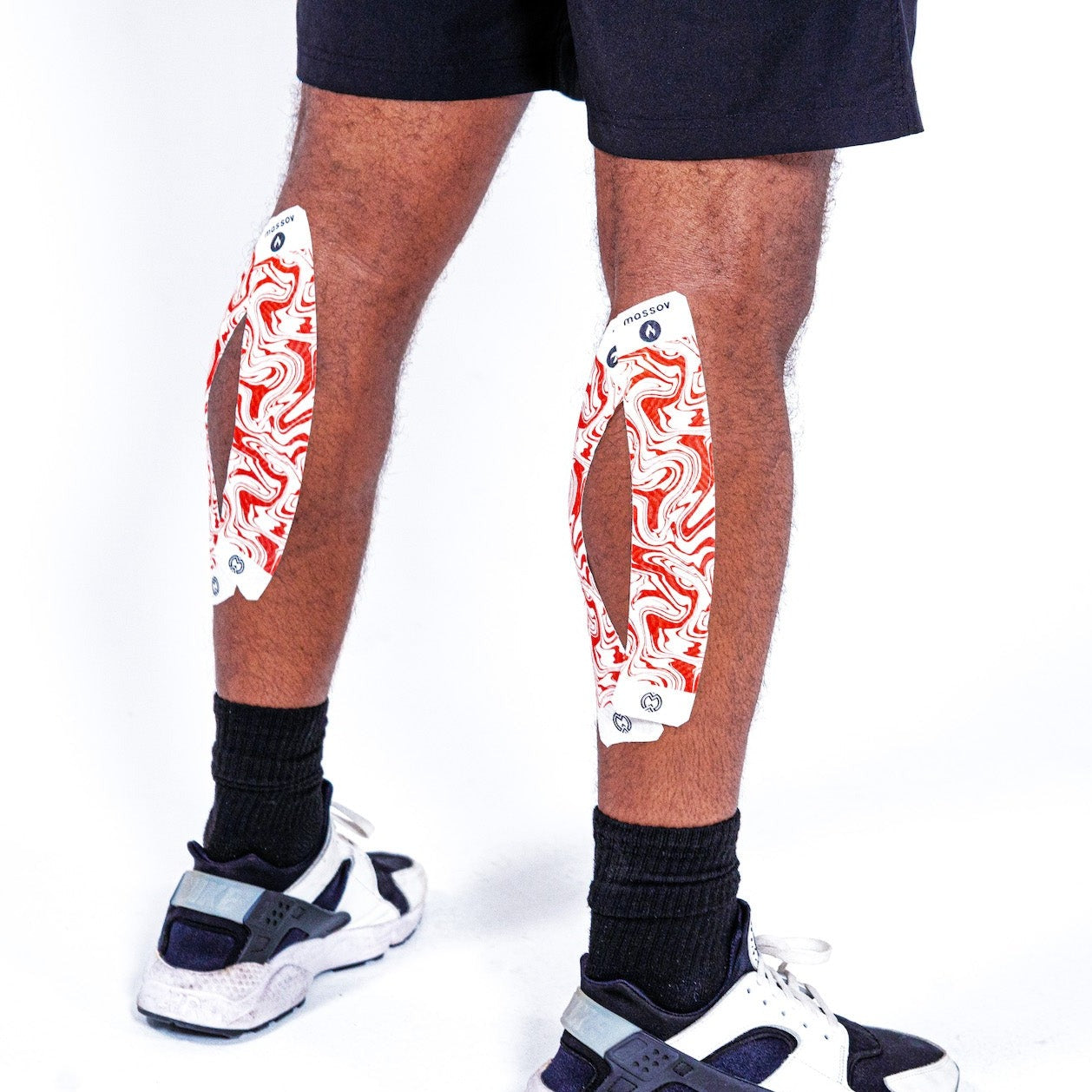 A person in black shorts, black socks, and white sneakers shows the back of their legs, where colorful Massov Heat Therapy Kinesiology Tape with red-and-white patterns and cutout designs is strapped to their calves.