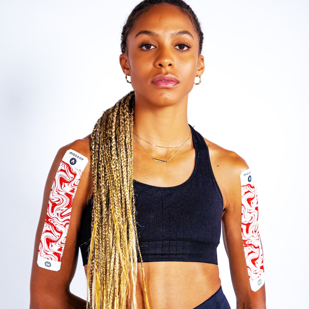 A woman with long, blonde box braids wears a black sports bra and Massov Heat Therapy Kinesiology Tape with a red and white swirl pattern on both upper arms, posing against a plain white background.