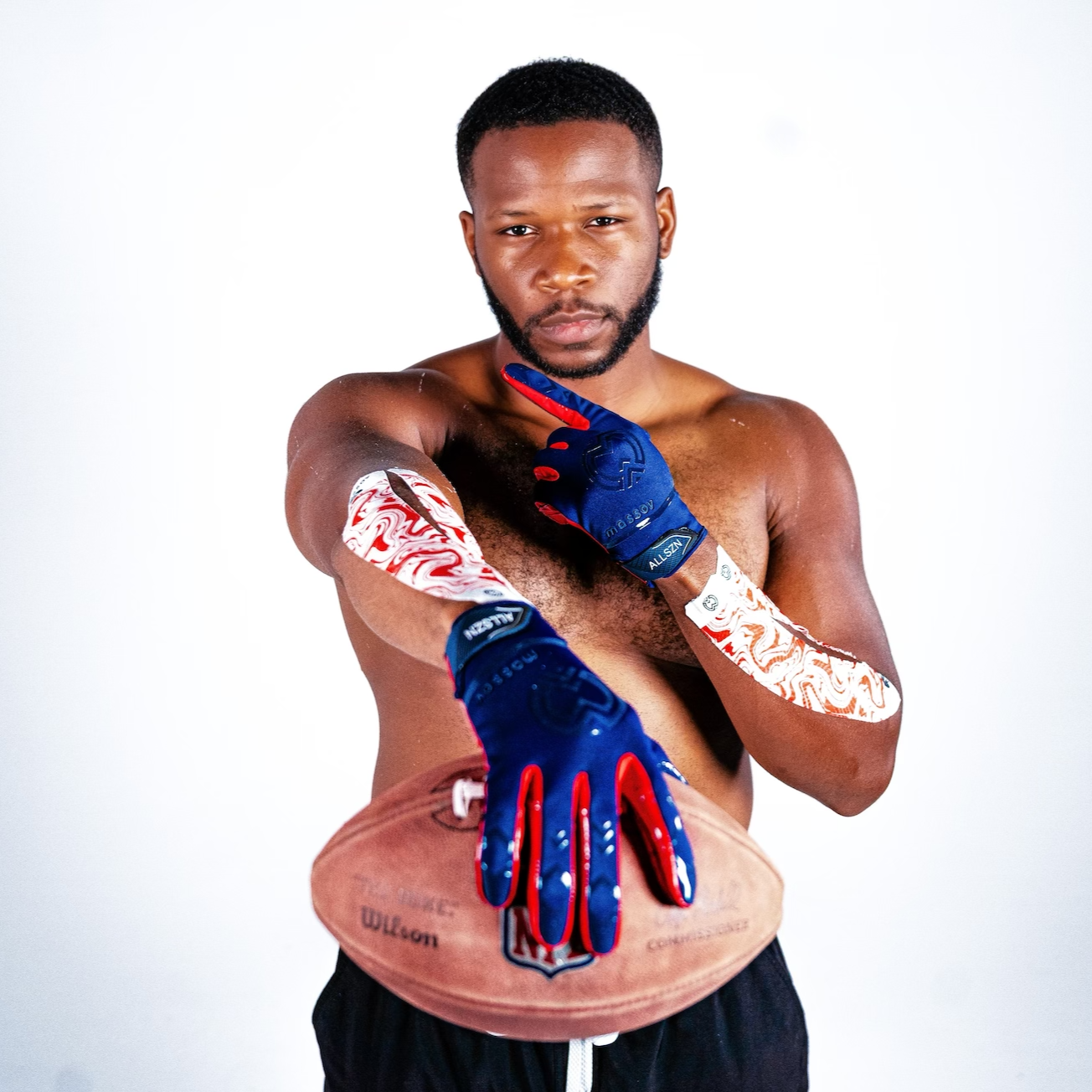 A shirtless man with short hair and arm tattoos wears blue football gloves and Massov Heat Therapy Kinesiology Tape, holding a Wilson NFL football while pointing forward. He stands against a plain white background, looking at the camera.