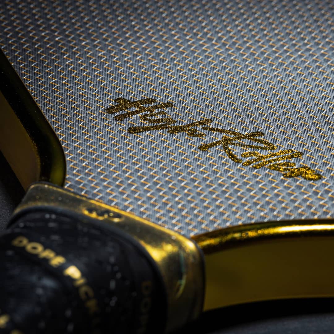 Close-up of the Dope Pickleball Co. High Roller Pickleball Paddle—The Original Chrome Gold. Features a textured gray and gold surface with golden Asian script, black grip tape with gold lettering, and a sleek gold edge.