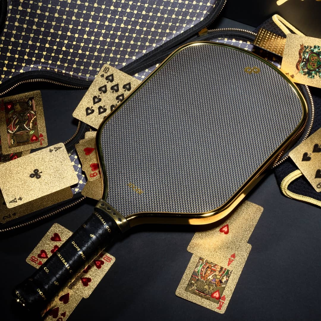 The High Roller Pickleball Paddle by Dope Pickleball Co. shines in chrome gold amid dazzling cards—spades, hearts, and kings—on a sleek case, merging luxury sports gear with stylish gaming vibes.