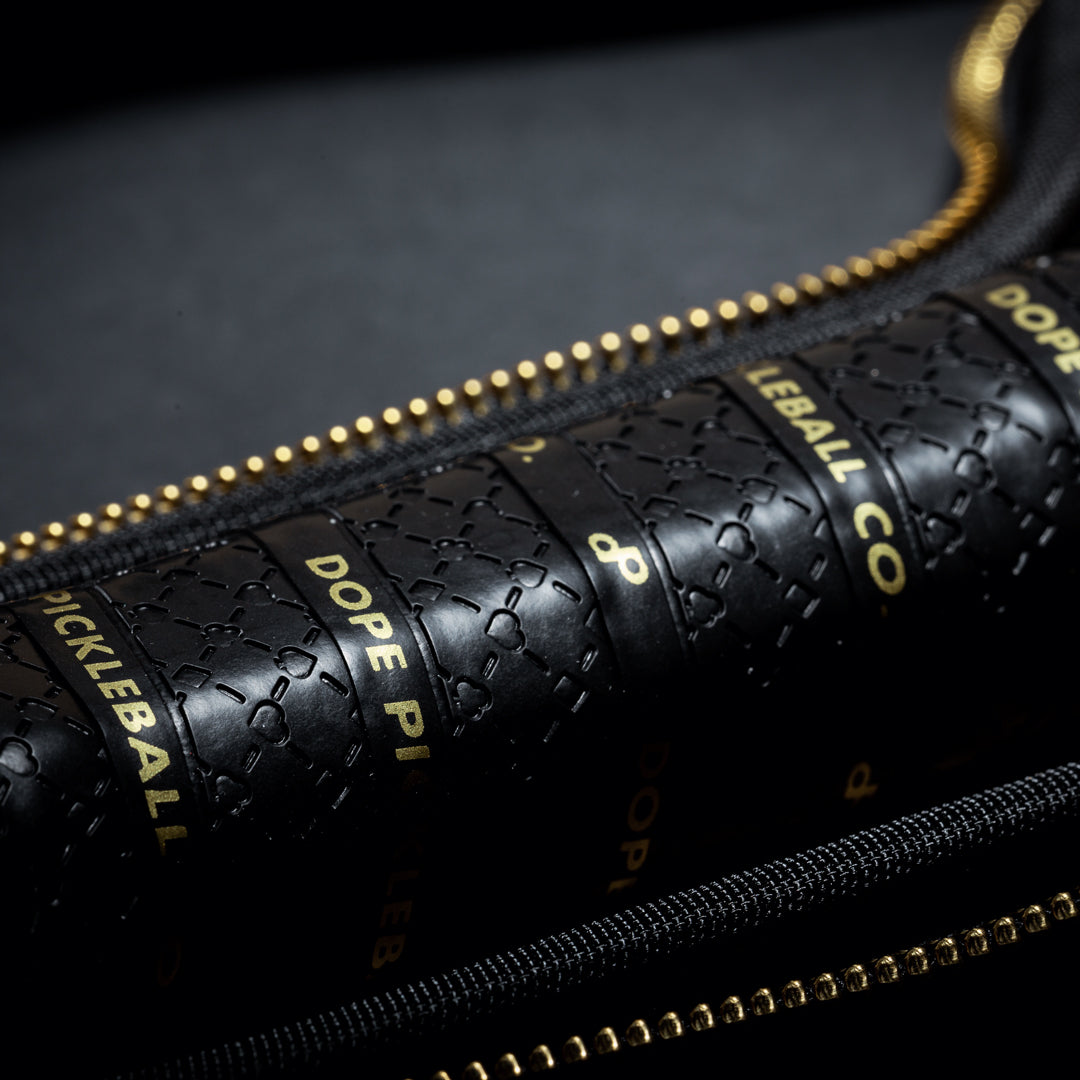 Close-up of the High Roller Pickleball Paddle by Dope Pickleball Co.—a black carbon fiber handle with embossed patterns and gold "DOPE" text. The handle is partially covered by a black zipper with gold teeth, set against a dark background.