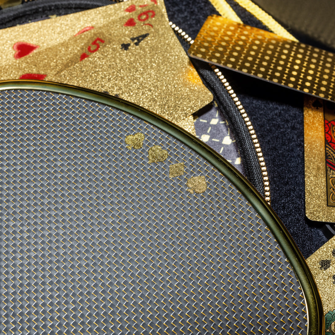 A close-up of glittery gold playing cards on a textured surface, with the Dope Pickleball Co. High Roller Pickleball Paddle - The Original Chrome Gold in front, its circular mesh pattern reflecting gold and blue tones for a luxurious look.