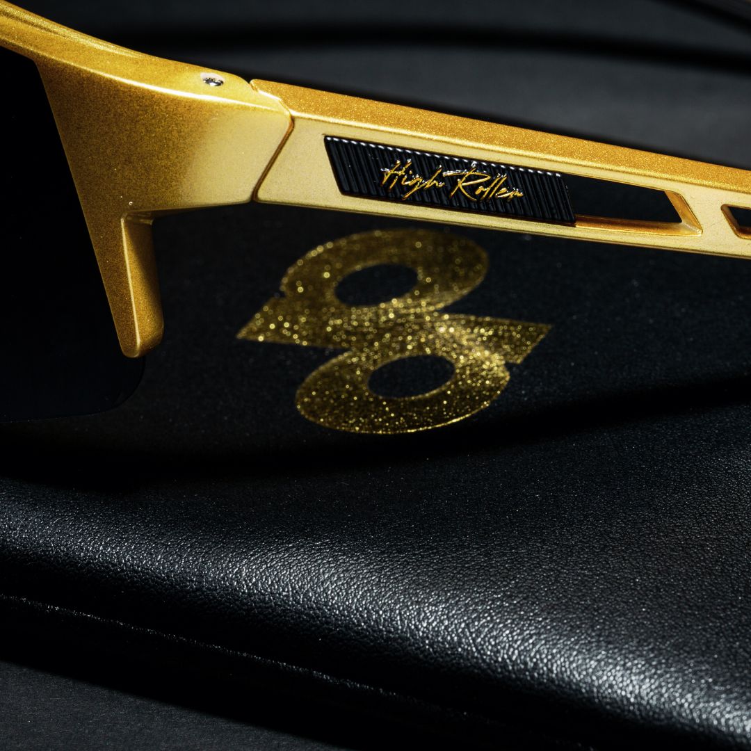 Close-up of Dope Pickleball Co. High Roller Performance Pickleball Sunglasses in gold with black lenses on black leather. The frame reads "High Roller" in cursive, and a glittery gold 8 is subtly reflected beneath the sunglasses.