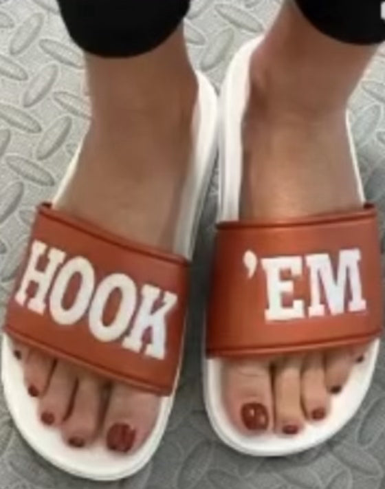 A person stands on patterned gray flooring wearing Runway Athletics HOOK 'EM Slides - LIMITED EDITION, featuring wide brown straps with "HOOK" on the left and "'EM" on the right in bold white letters. Their toenails are painted dark red.