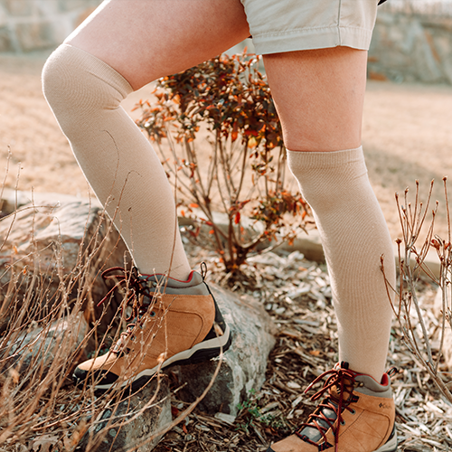 Wearing iReliev Bamboo Anti-Fatigue Compression Socks with khaki shorts and brown hiking boots, a person stands outdoors on rocky ground amid dry grass, one foot on a rock as sunlight casts warm tones over the scene.