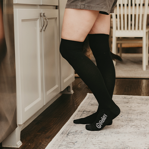 A person wearing iReliev’s Compression Therapy Bundle black knee-high socks with “@thekelly” in white on the toes stands on a light rug in a kitchen by white cabinets; only their legs and shorts are shown, with a wooden chair behind them.