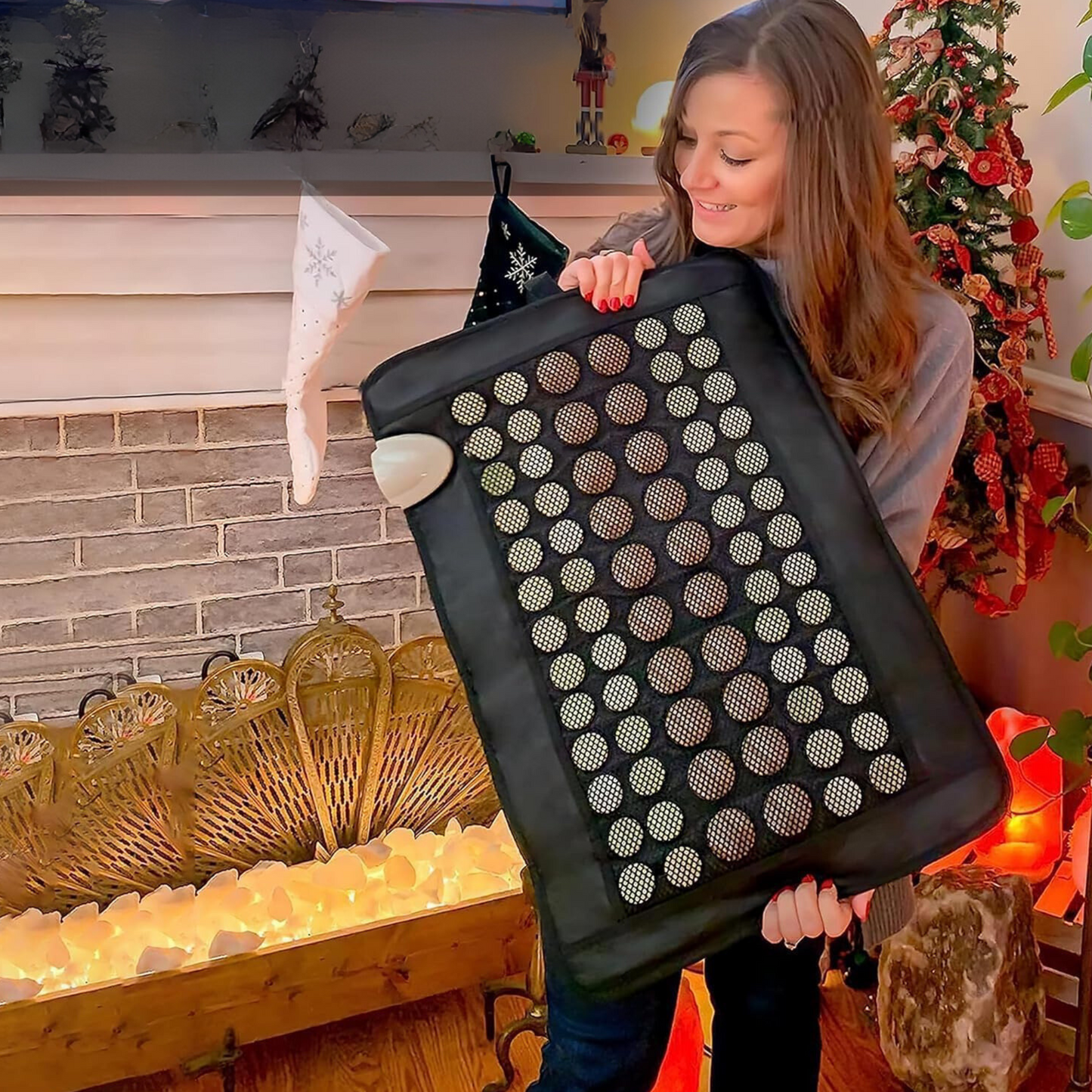 A woman with long brown hair, wearing a gray sweater, smiles while holding the iReliev Far Infrared Natural Jade and Tourmaline Heating Pad (23.5" x 16") in a cozy living room decorated for Christmas with a lit salt lamp and brick fireplace.