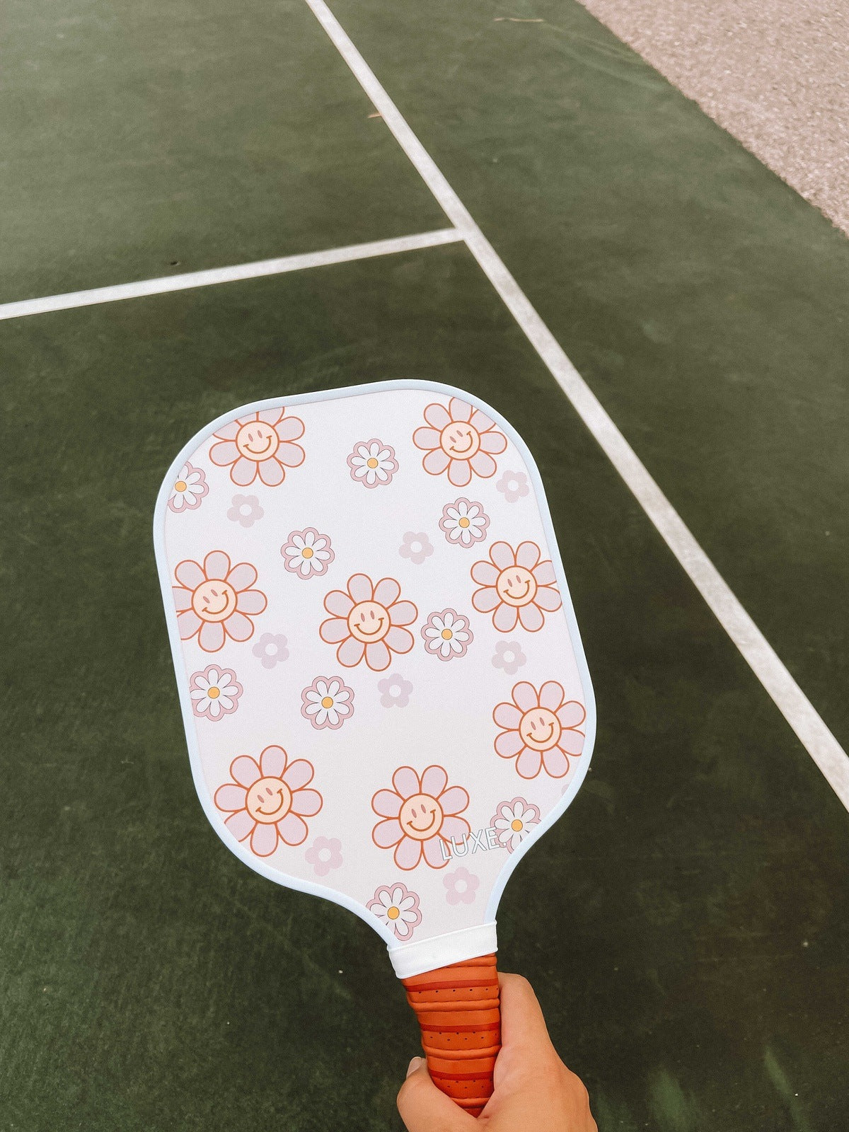 A hand holds the LUXE Pickleball Smiley Flower paddle—USA Pickleball Approved, featuring pink and orange smiling flowers—on a green court with white lines, making it ideal for beginners.