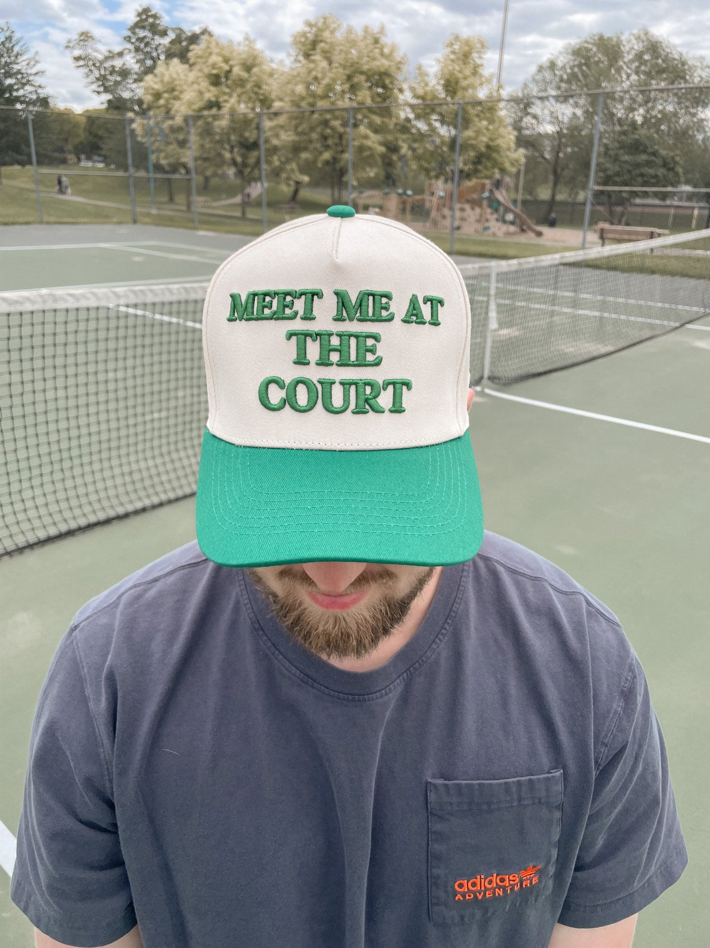 A bearded individual stands on a tennis court wearing the LUXE Pickleball "Meet Me At The Court Hat," a green-and-white snapback with bold green text. They pair it with a blue Adidas t-shirt; the net, trees, and playground are in view.