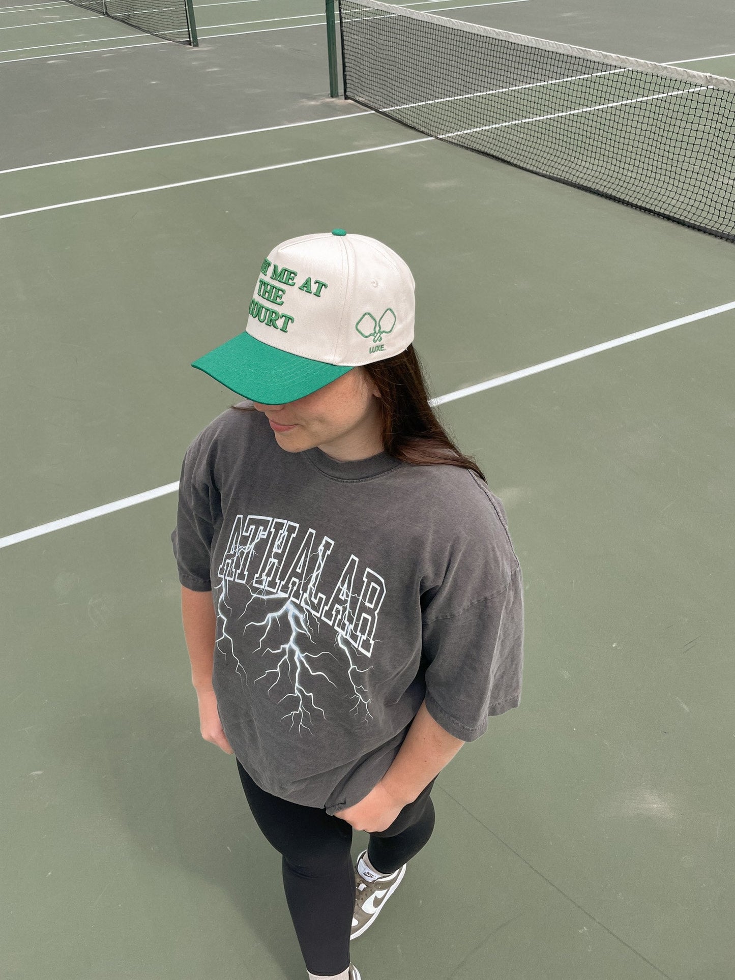 A woman stands on a tennis court, wearing the Meet Me At The Court Hat by LUXE Pickleball for stylish sun protection.