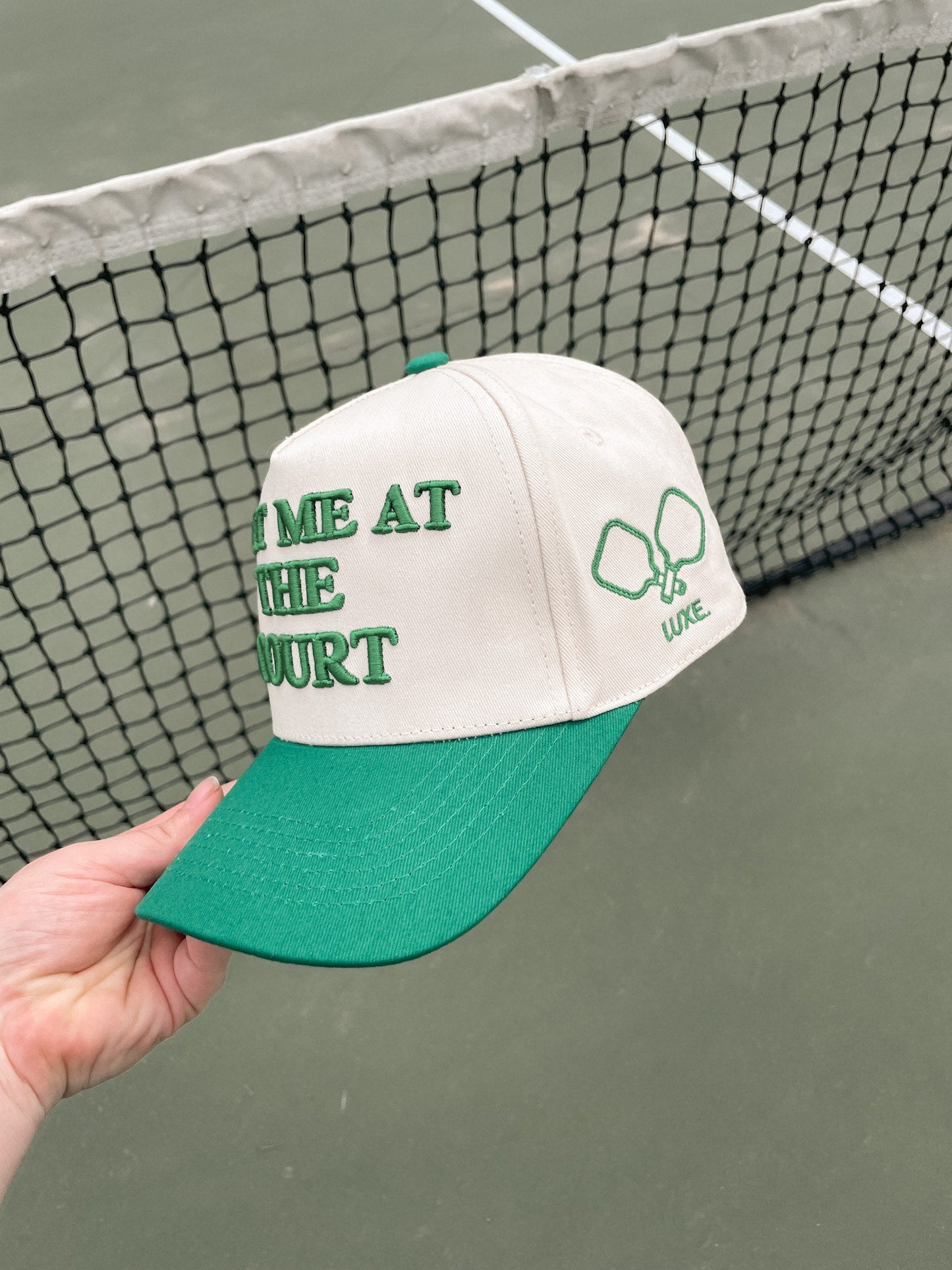 A hand holds the LUXE Pickleball Meet Me At The Court Hat—a white and green snapback with "MEET ME AT THE COURT" in green, plus two crossed tennis rackets and "LIKE" embroidered—shown in front of a tennis net on an outdoor court.