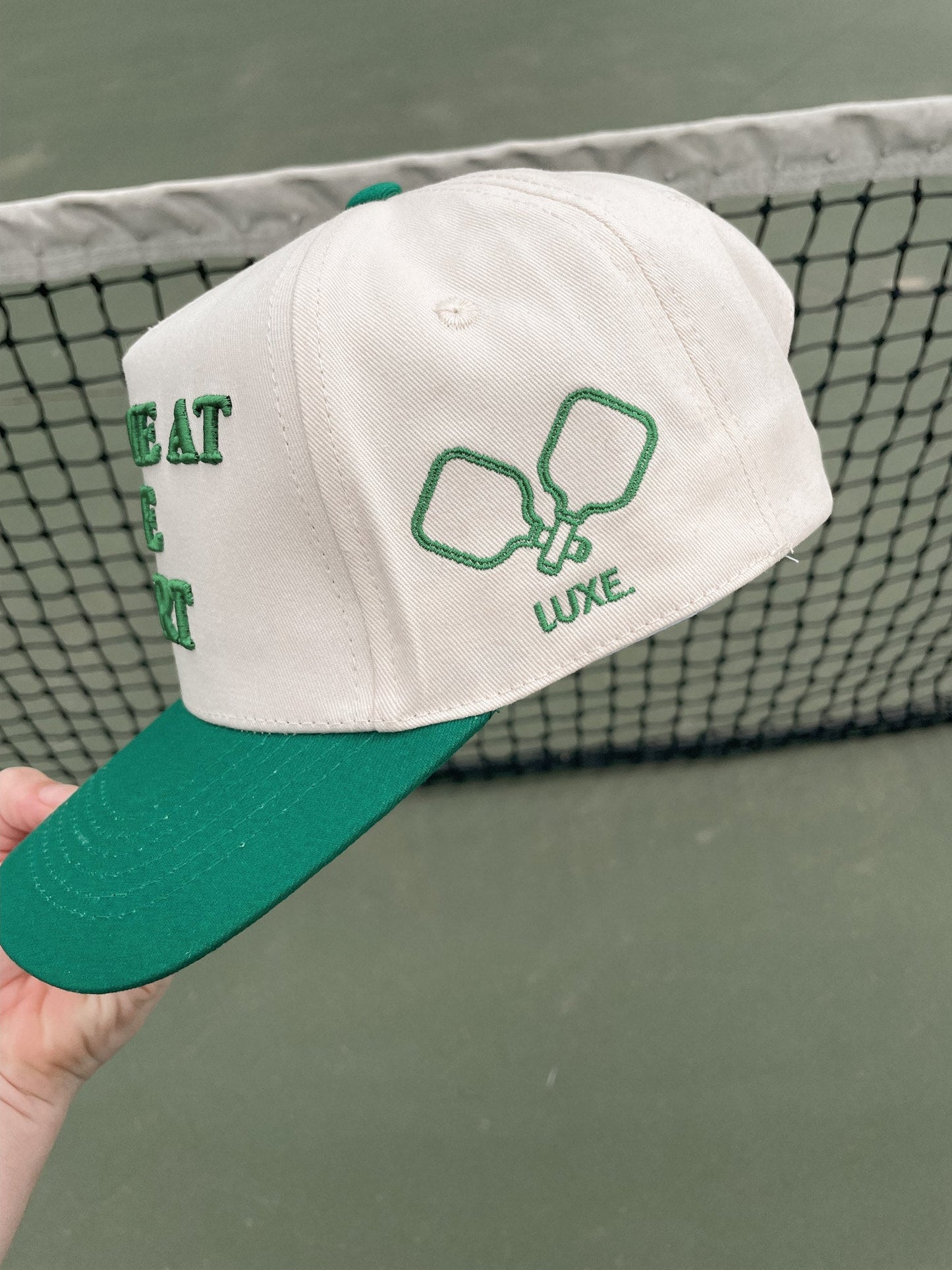 A hand holding the Meet Me At The Court Hat by LUXE Pickleball, a stylish snapback perfect for sun protection.
