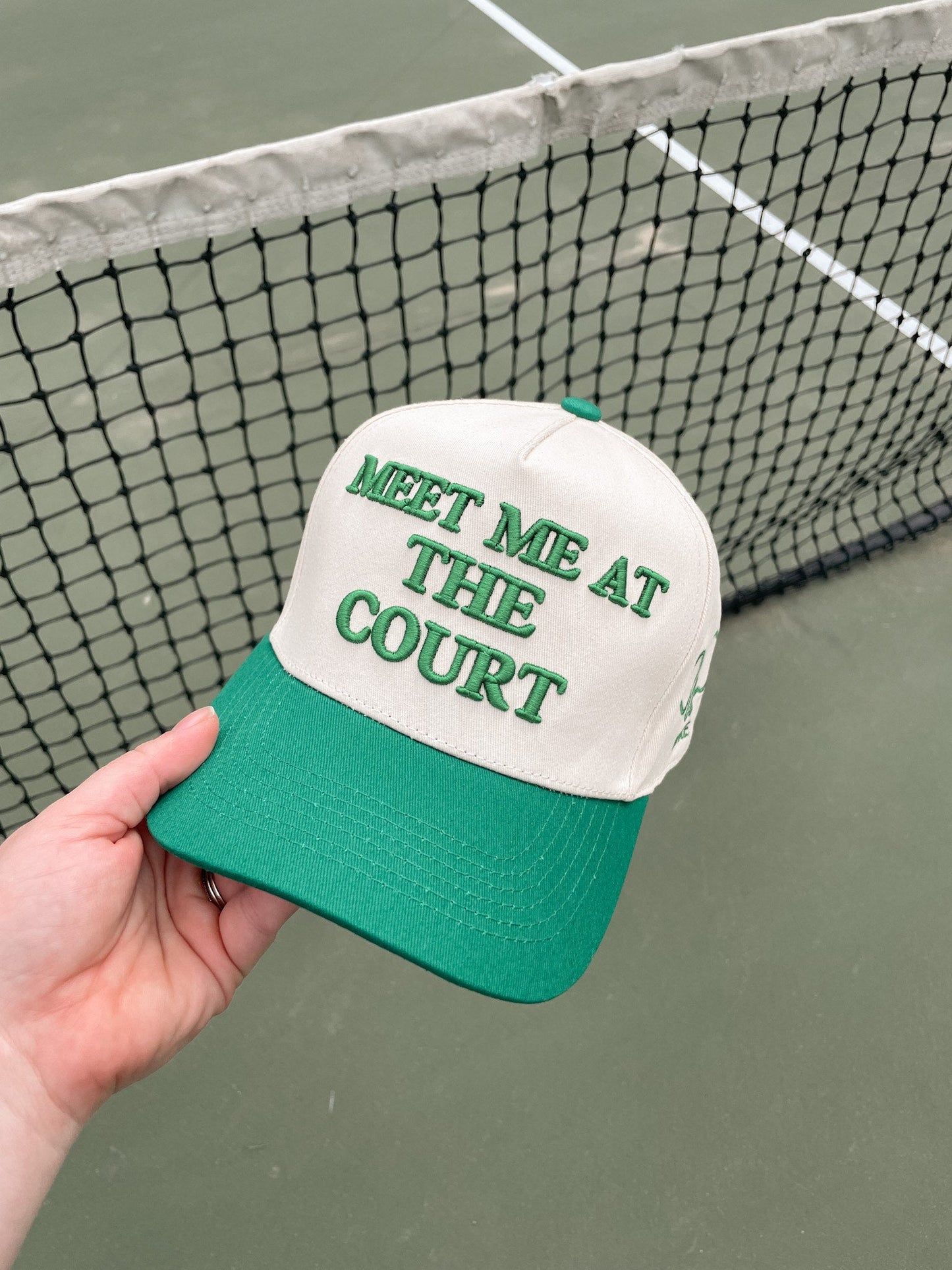 A hand holds the LUXE Pickleball Meet Me At The Court Hat, a white and green snapback featuring green embroidered text. Set against a tennis net and court, it offers both style and sun protection for your next match.