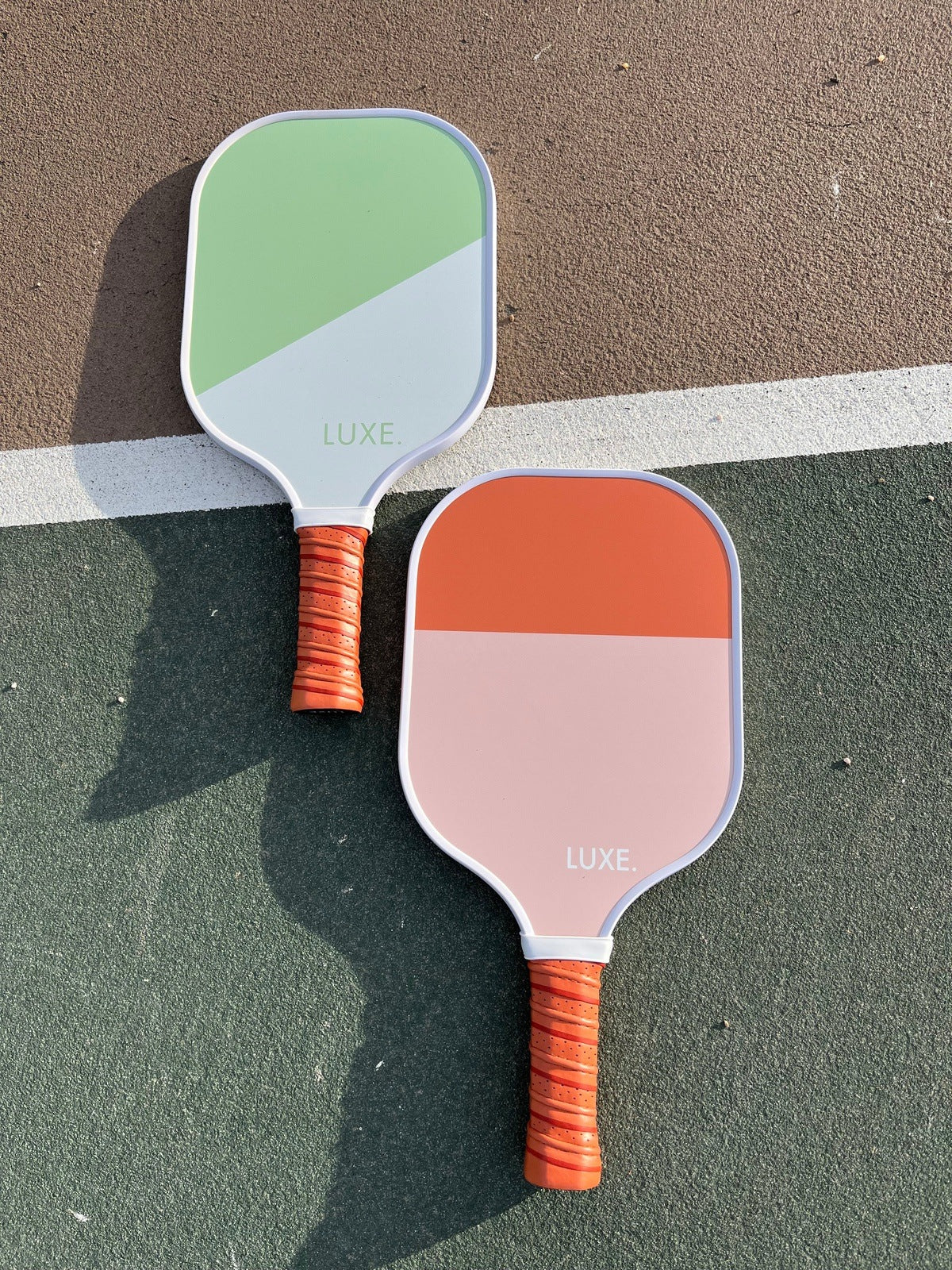 Two "Dipped - Leisure" paddles by LUXE Pickleball lie on an outdoor court. The paddles have fiberglass surfaces and orange-striped grips, casting shadows on the green and brown court separated by a white line.