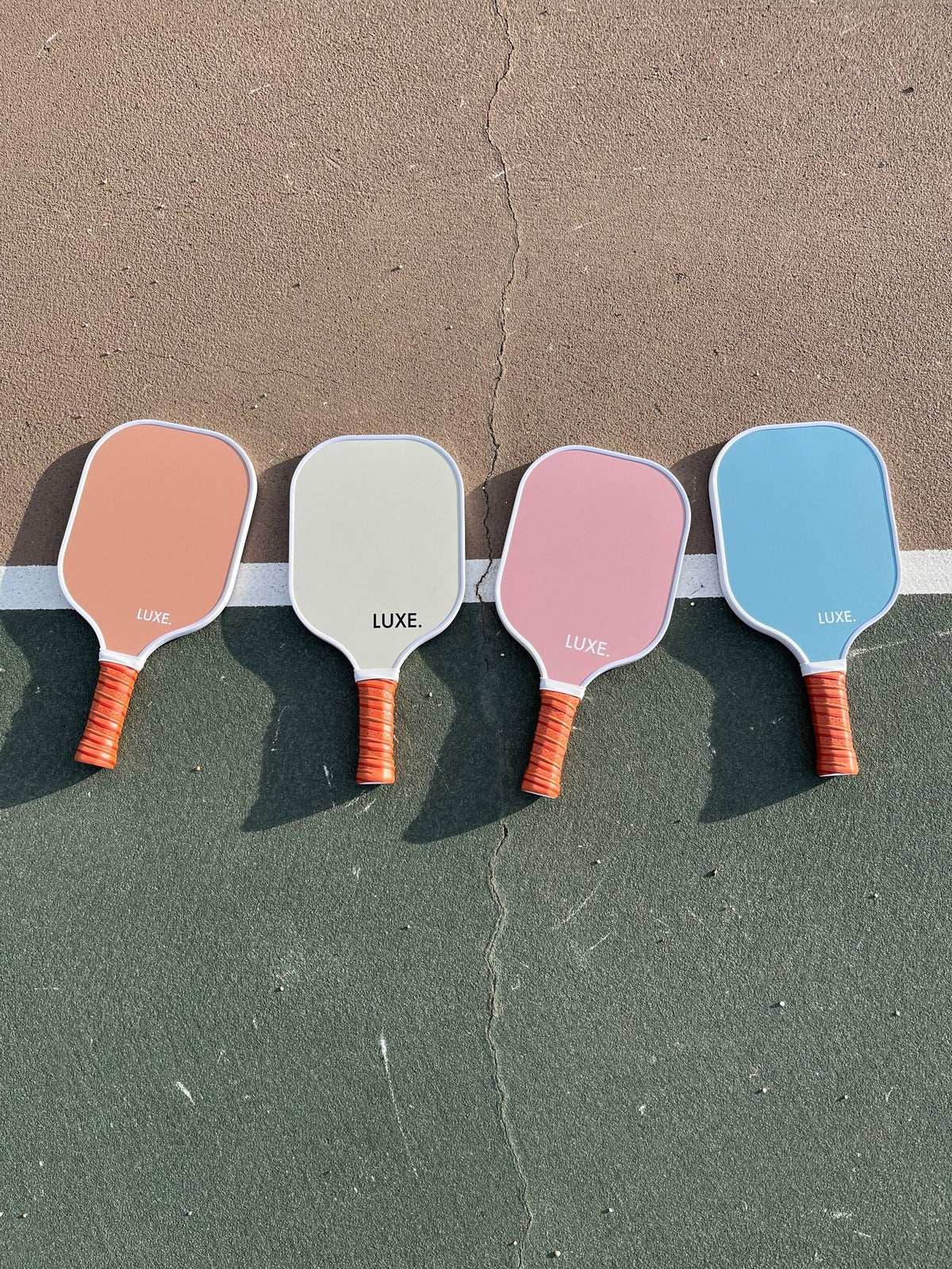 Four LUXE Pickleball Aesthetic Solids - Ace paddles with red-orange handles and pastel faces (tan, light gray, pink, and light blue) are lined up on an outdoor court across a white line between tan and green surfaces.