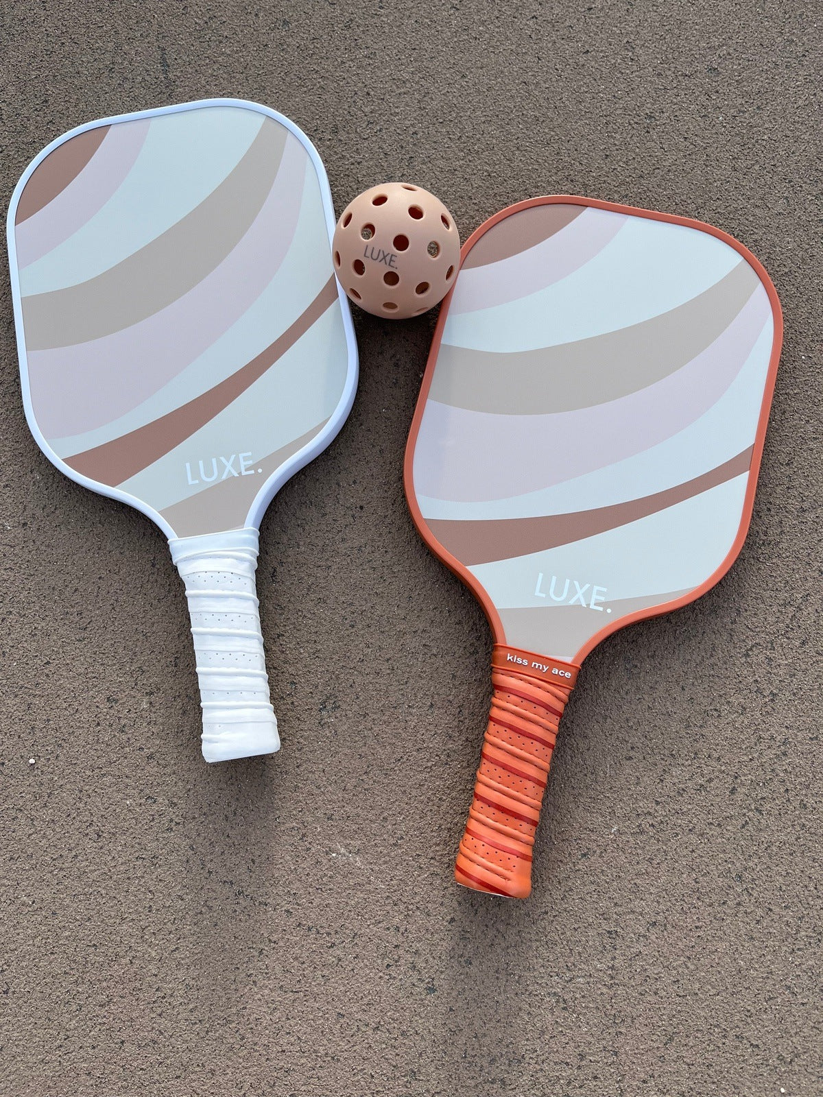 Two LUXE Pickleball paddles from the Pink and Rust set—one pink, one rust—lie on a textured outdoor court with a perforated orange pickleball placed between them. All items are branded LUXE Pickleball.