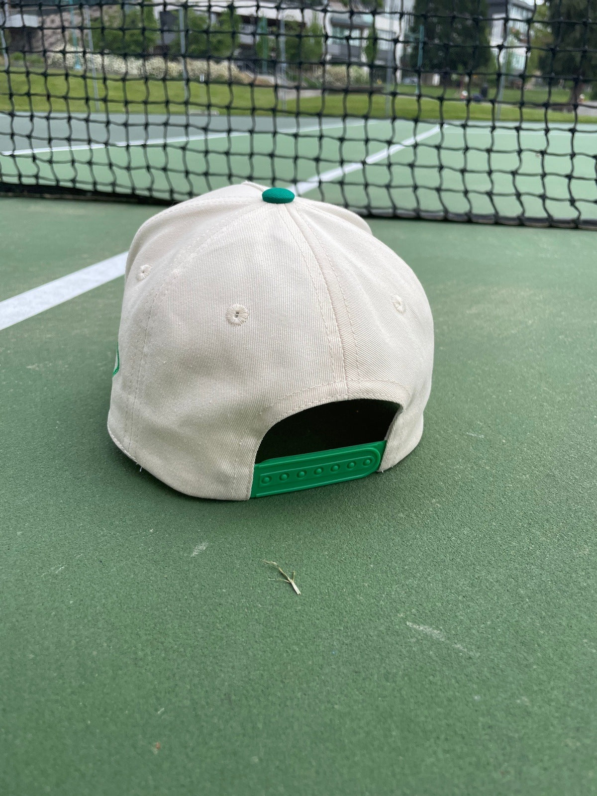 The LUXE Pickleball Meet Me At The Court Hat, in beige with a green adjustable strap, sits upside down near the net on a green tennis court with white lines and a black net, surrounded by blurred grass and buildings.