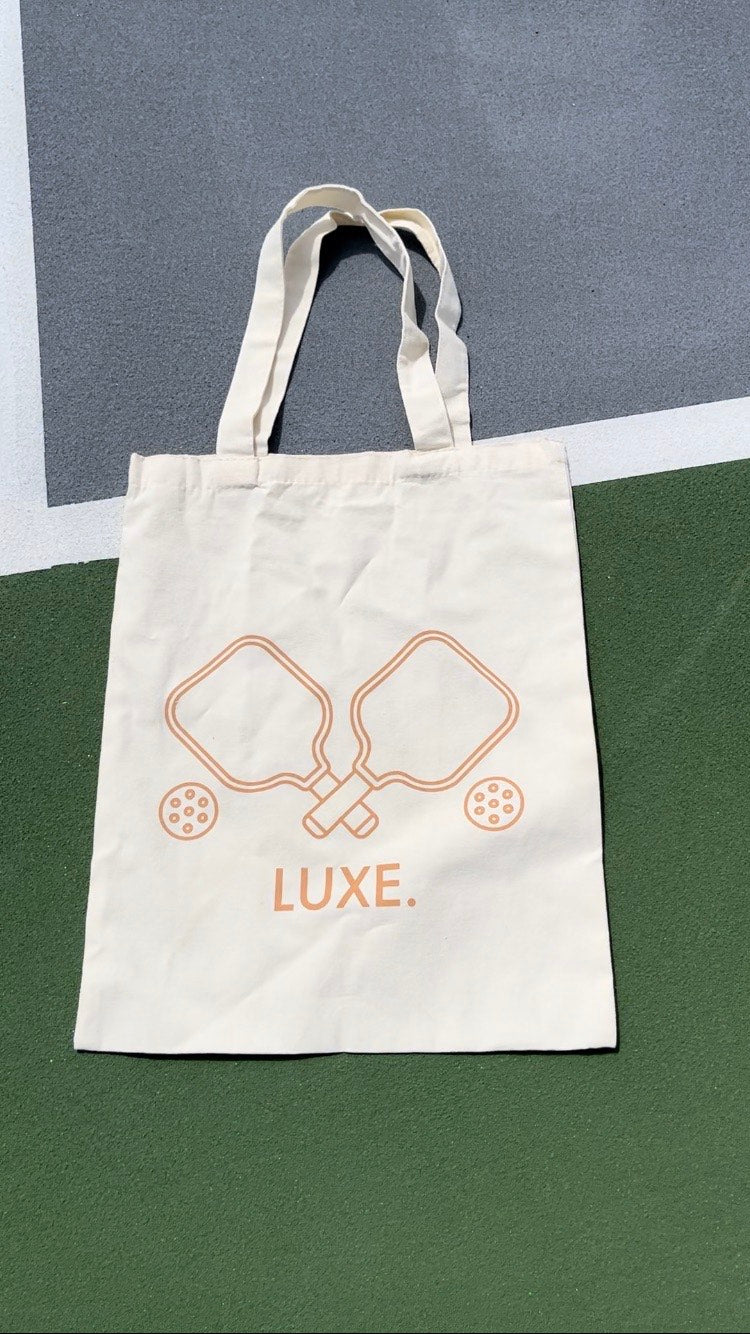 A cream-colored LUXE Pickleball Tote Bag with two handles rests on a green, gray, and white surface. It features a minimalist orange outline of crossed paddles and pickleballs above the bold LUXE. brand name.