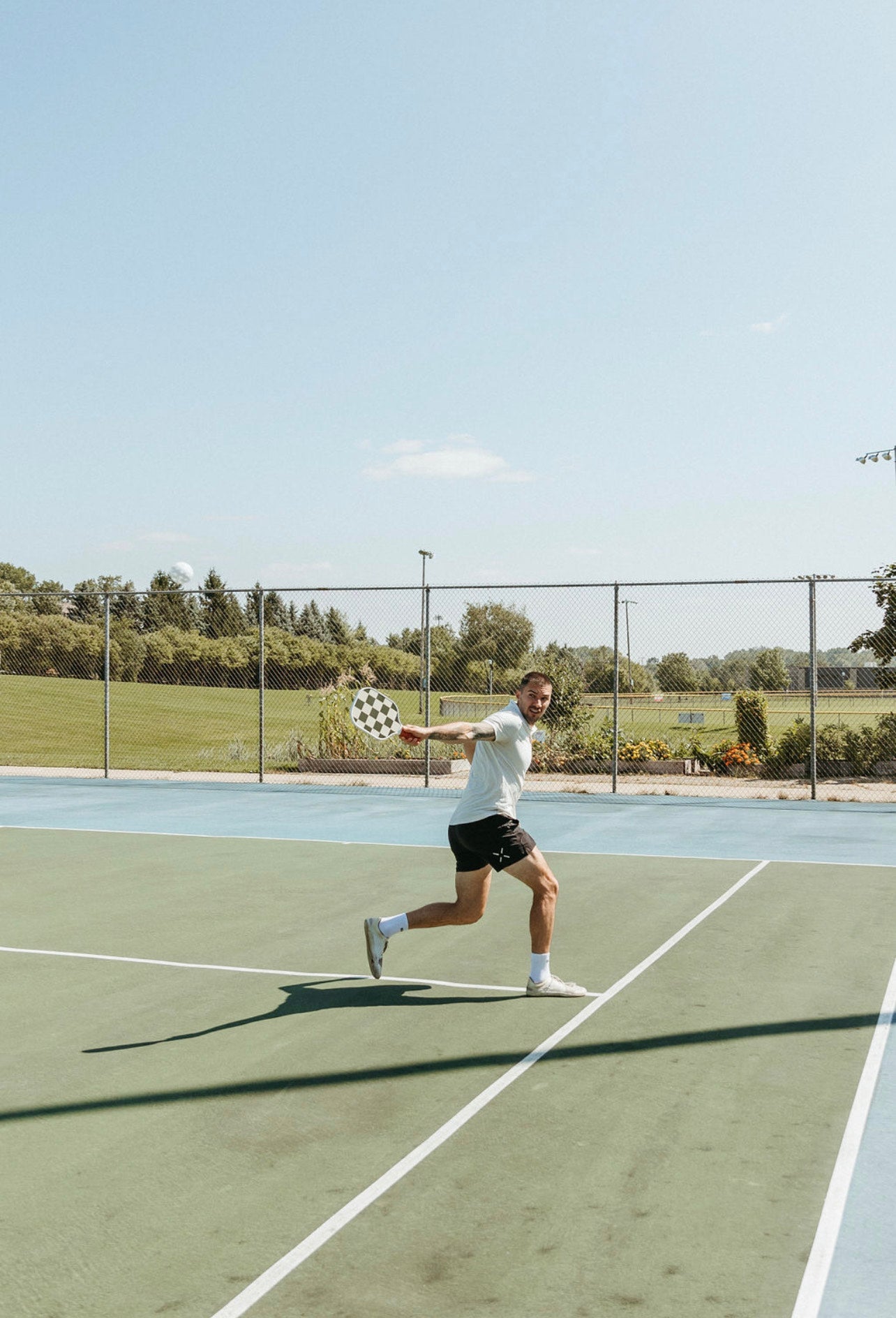 A man in a white shirt and black shorts plays pickleball on an outdoor court, swinging the LUXE Pickleball Checkmate paddle. Behind him are a fence, grassy field, trees, and a clear blue sky.