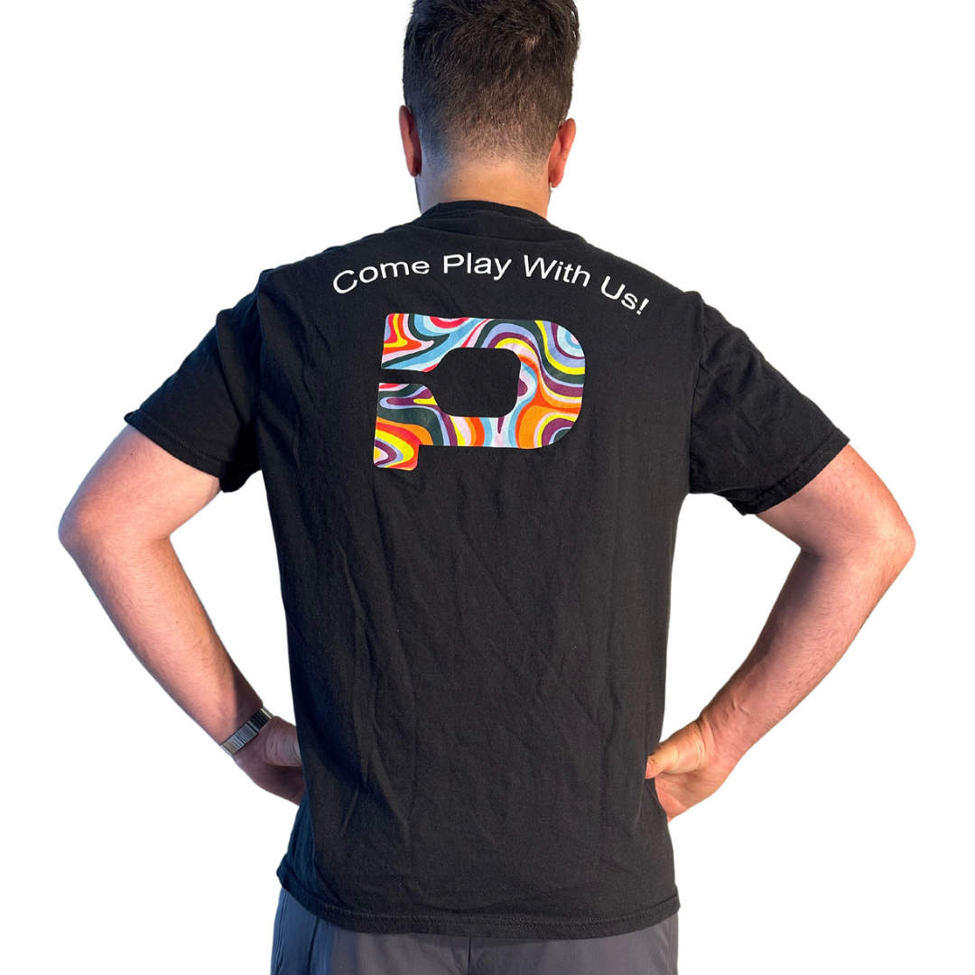 A person faces away from the camera, wearing Playly Store's unisex black cotton "Playly - Logo T," which showcases a colorful abstract square design with "Come Play With Us!" in white above the pattern.