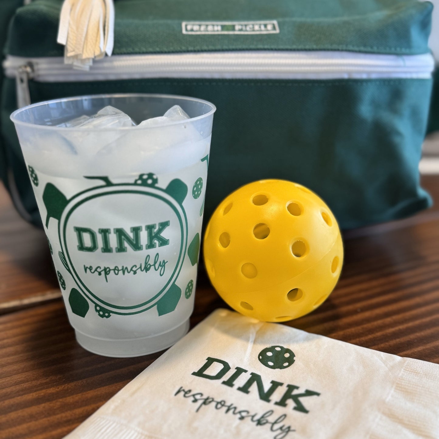 A plastic cup of ice water with “DINK responsibly” and pickleball motifs sits by a yellow pickleball and Fresh Pickle’s Pickleball Cocktail Napkins, all on a wooden surface. A green bag labeled “OPEN PICKLE” completes the playful scene.