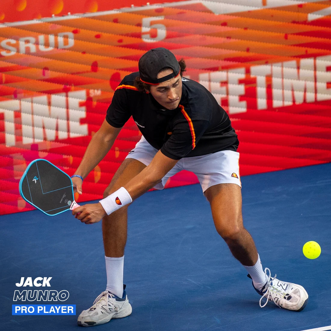 A young male pickleball player in a black shirt, white shorts, and a backward cap lunges low on an indoor blue court to hit a yellow ball with his Hesacore Grip Pickleball Gel Grip paddle (5.25" soft feel, light blue). Red walls are in the background.