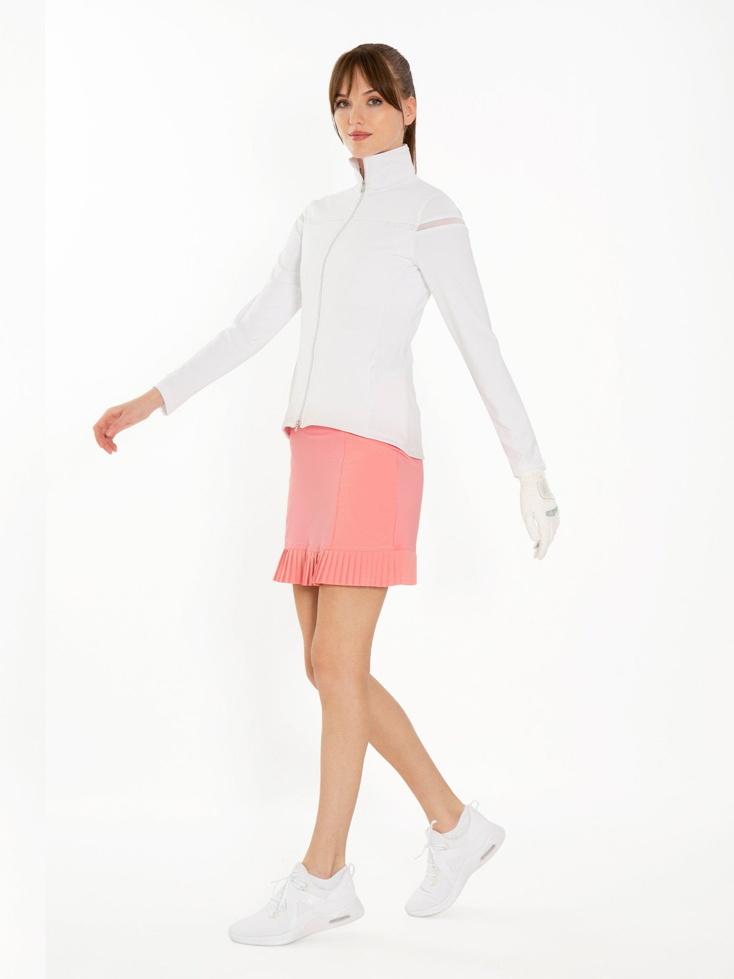 A woman poses confidently against a white background wearing the inPhorm NYC Kai 17" Pleated Skirt in Plumeria, paired with a white zip-up jacket, white athletic shoes, and a glove on her left hand.