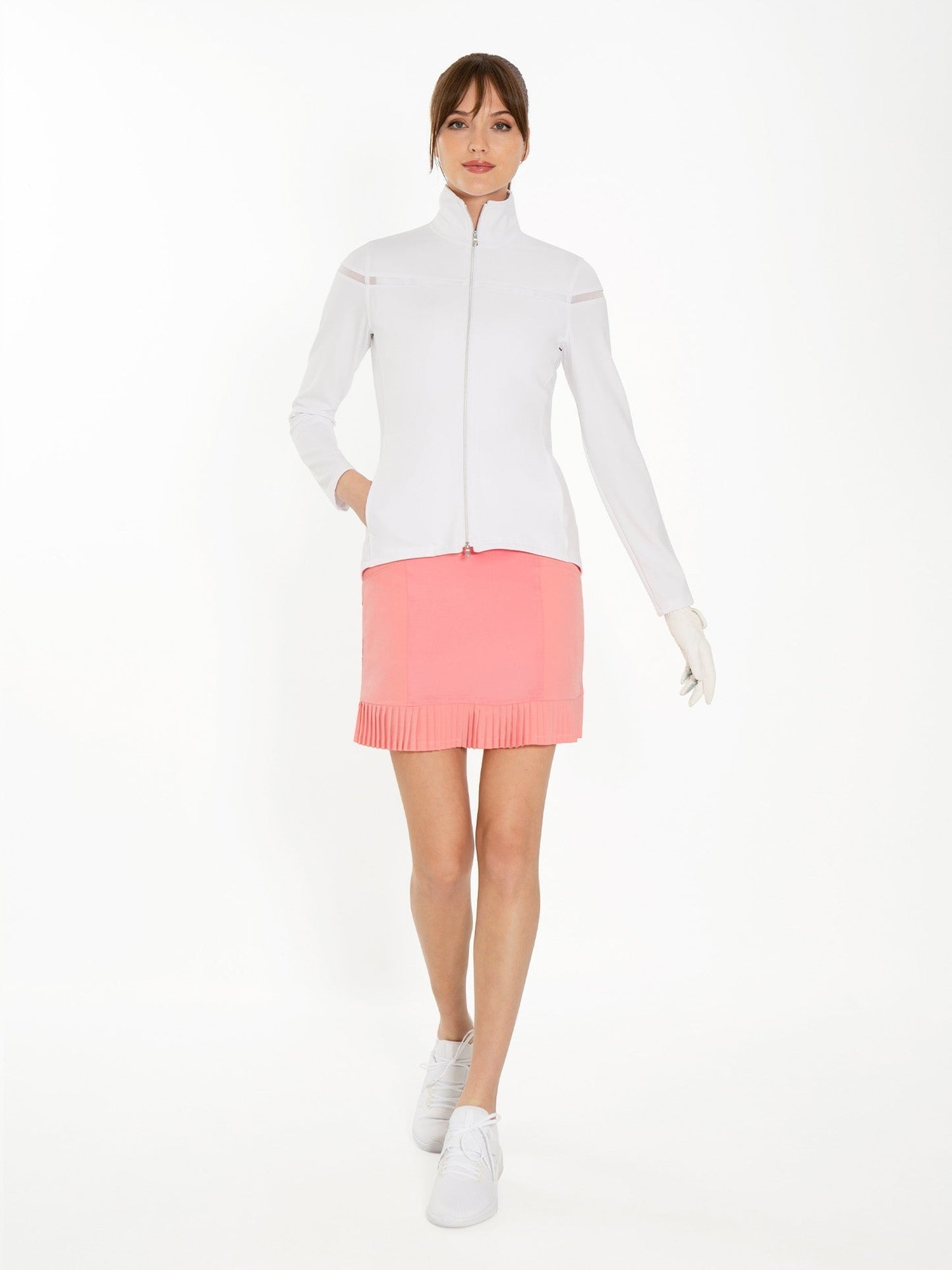 A woman stands against a white background wearing a white zip-up jacket, the inPhorm NYC Kai 17" Pleated Skirt - Plumeria, white sneakers, and a single white golf glove. Her brown hair is tied back with bangs; her left hand rests in her skirt pocket.