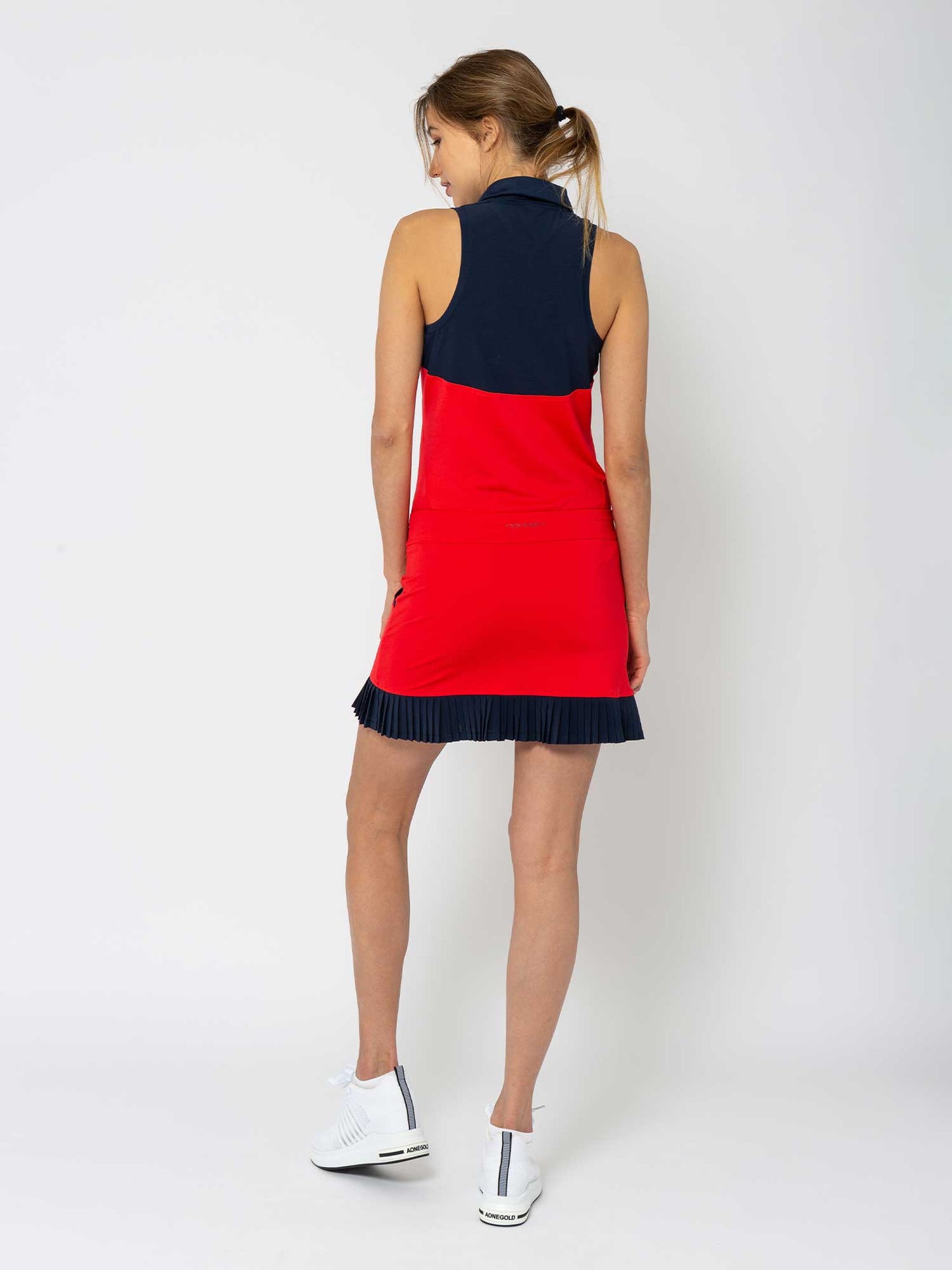 A woman with her brown hair in a ponytail faces away, wearing the inPhorm NYC Kai 17" Pleated Skirt in Poppy/Midnight and white athletic shoes, against a plain white background.