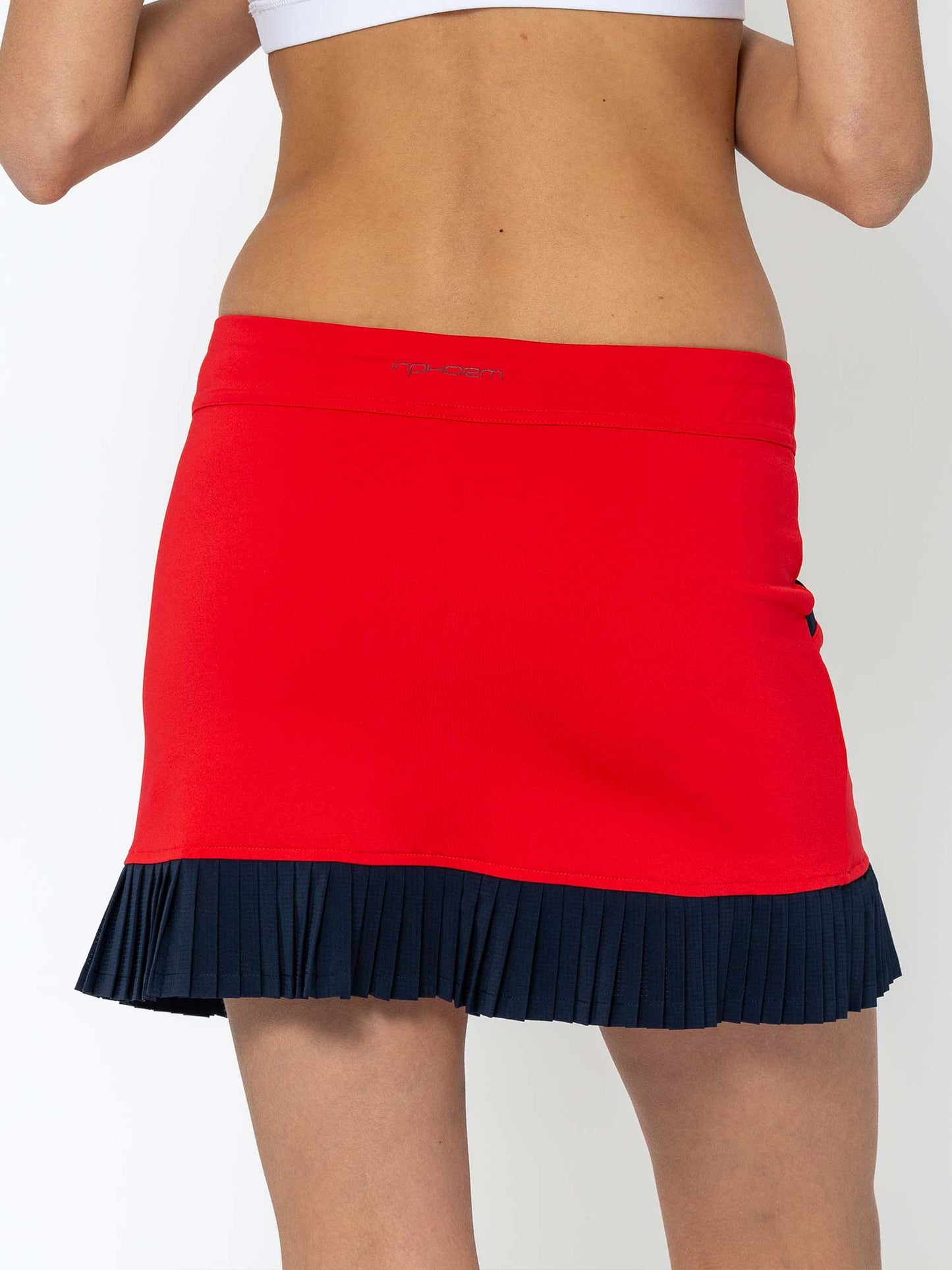 A person is shown from the back wearing the inPhorm NYC Kai 17" Pleated Skirt in Poppy/Midnight with dark pleated trim at the waist, paired with a white sports bra, revealing lower back and midriff. The background is plain white.