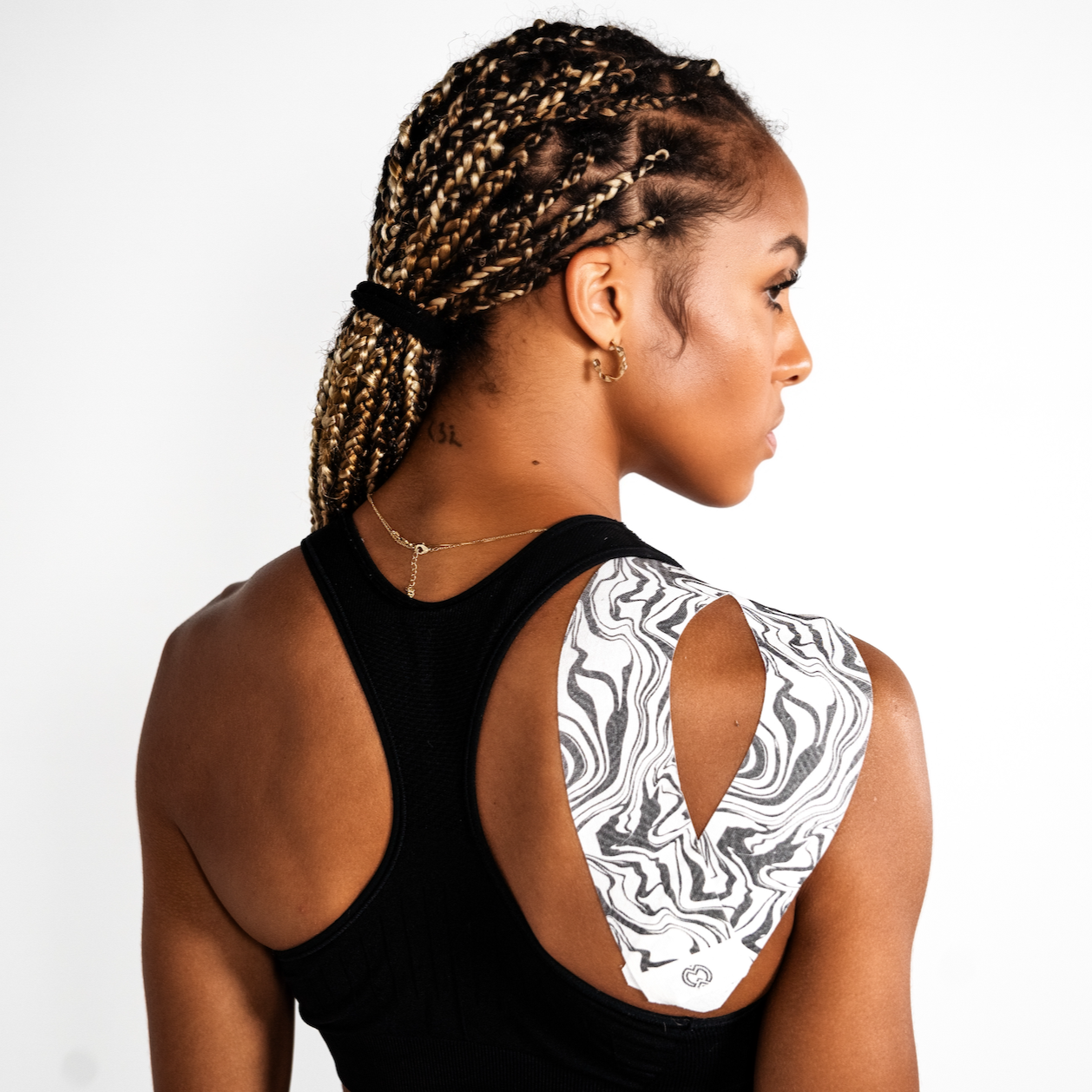 A woman with braided hair, in a black sleeveless top, faces away from the camera. Massov Kinesiology Tape in black and white covers her right shoulder, providing performance support against a plain white background.