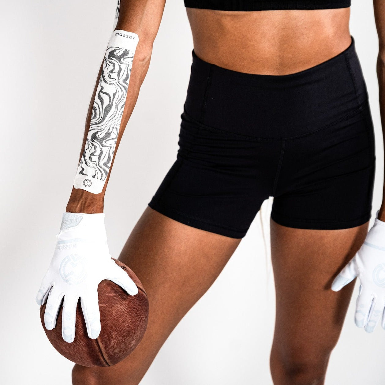 A person in black athletic shorts, a black sports top, and white gloves holds a brown football. Their left forearm is wrapped with Massov Kinesiology Tape featuring a black and white swirl pattern. The background is plain white.