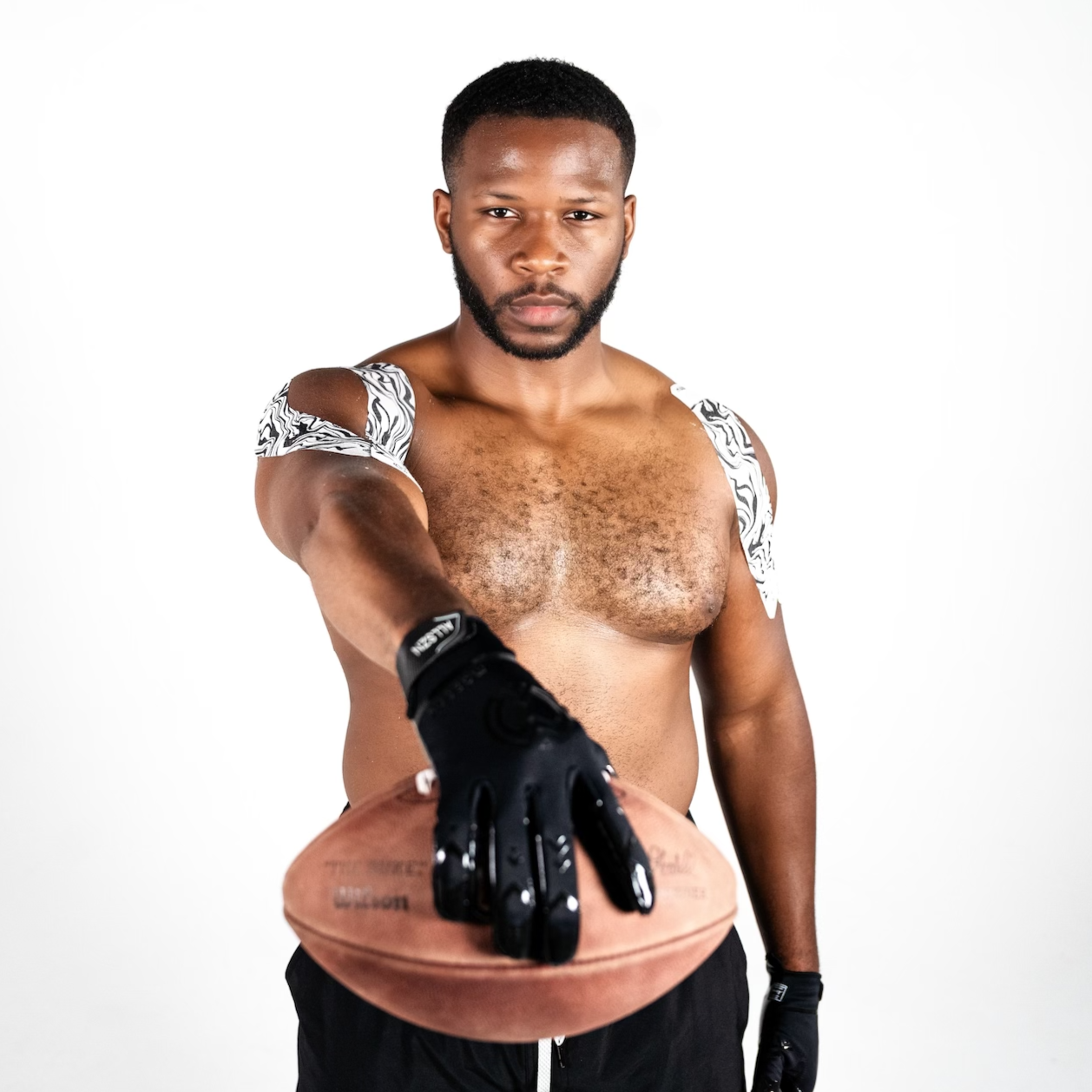 A muscular, shirtless man wears Massov Kinesiology Tape on his shoulders along with black gloves and shorts, holding an American football toward the camera. He stands against a white background, looking forward with a serious expression.