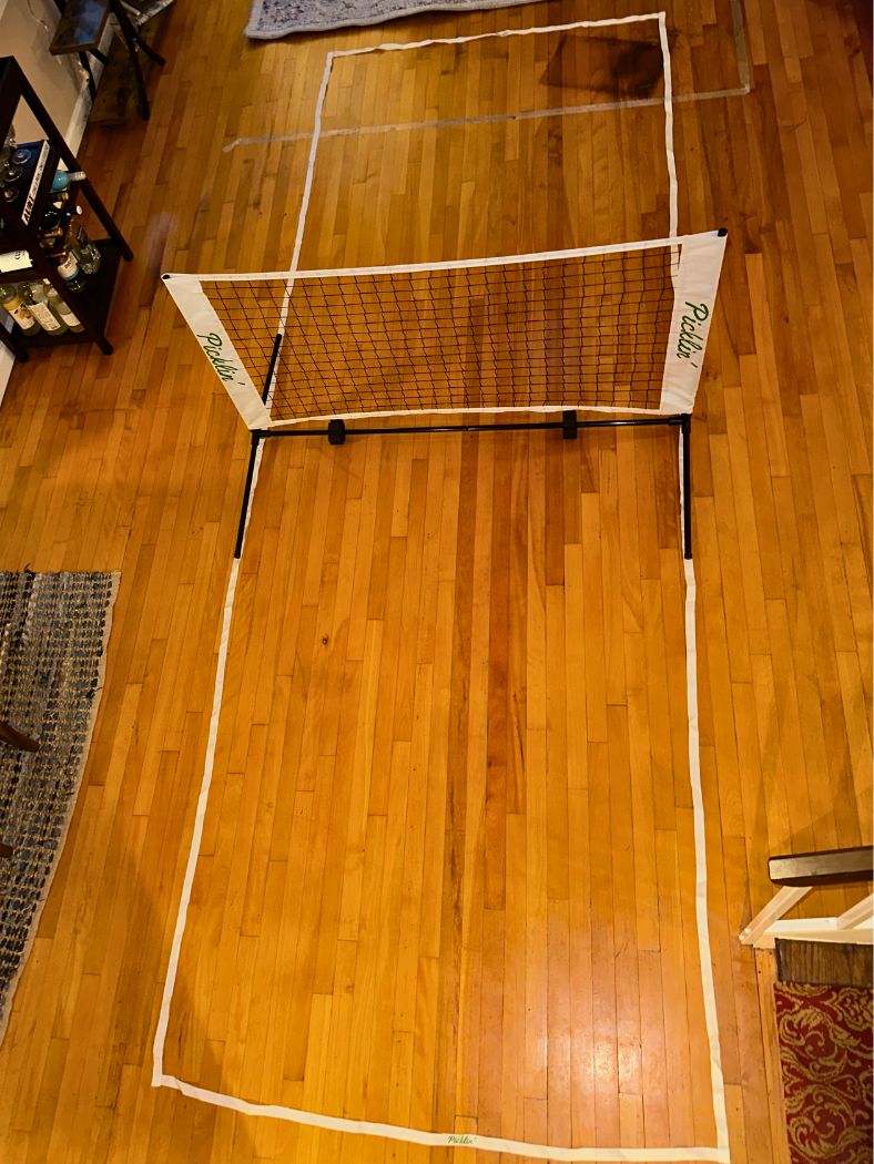 A Picklin™ Kitchen Lines (For Dink Net) mini pickleball court is set up indoors on a wooden floor with white tape marking the Kitchen Lines, the Dink Net across the width, and furniture and rugs around for convenient practice.