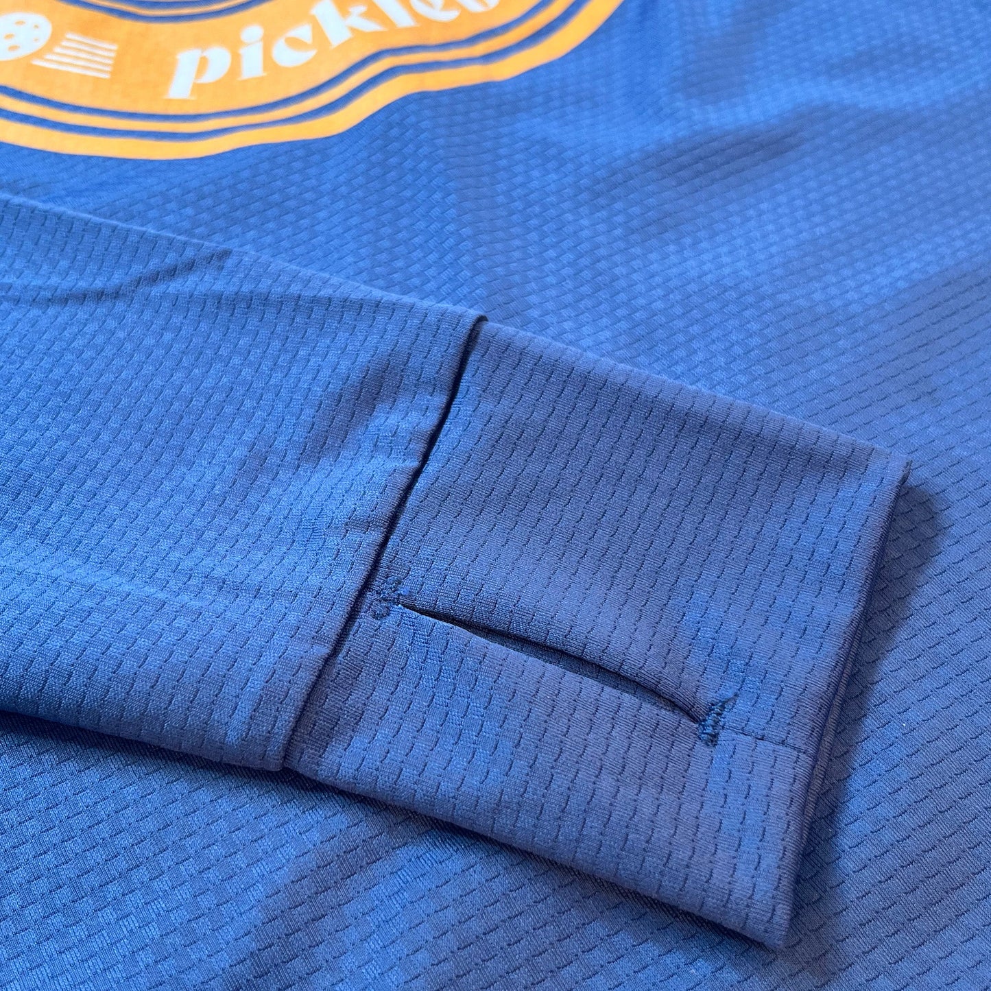 Close-up of the cuff on this lightweight, breathable Swinton Pickleball Navy Zip-Front Sun Hoodie features a thumbhole and subtle grid pattern. An orange and white “pickle” logo with partial pickleball adds an athletic touch.